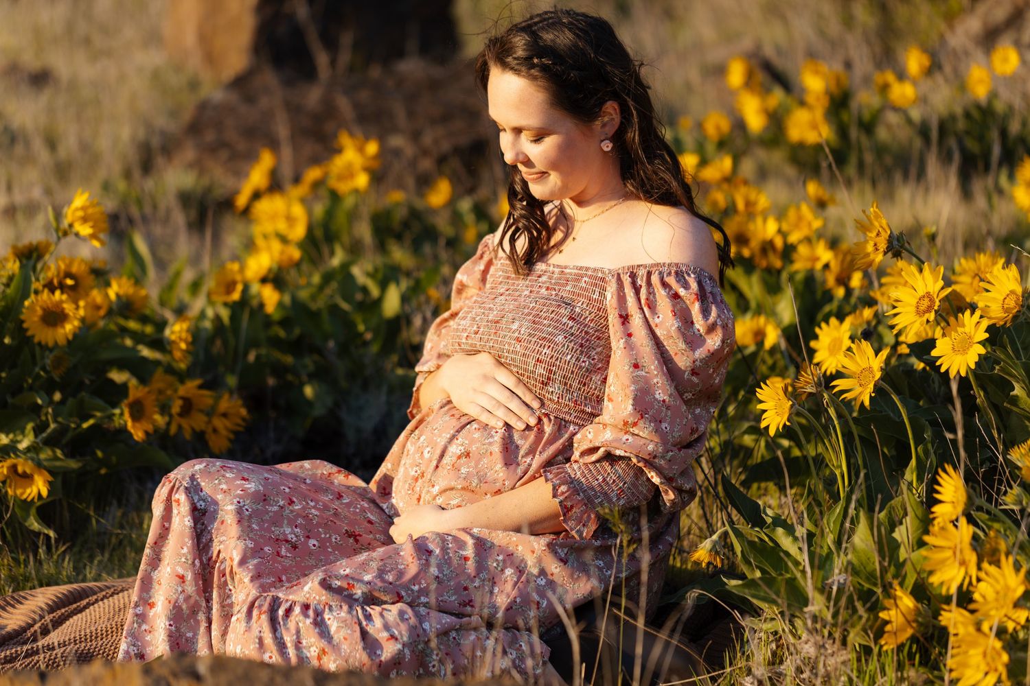 Expectant mother poses in floral dress surrounded by golden wildflowers.