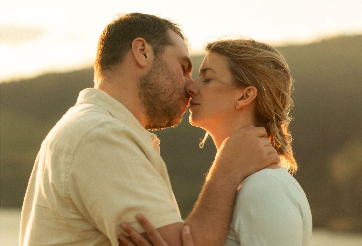 Romantic couple sharing a tender kiss outdoors at golden hour with a scenic lake and mountains in the background.