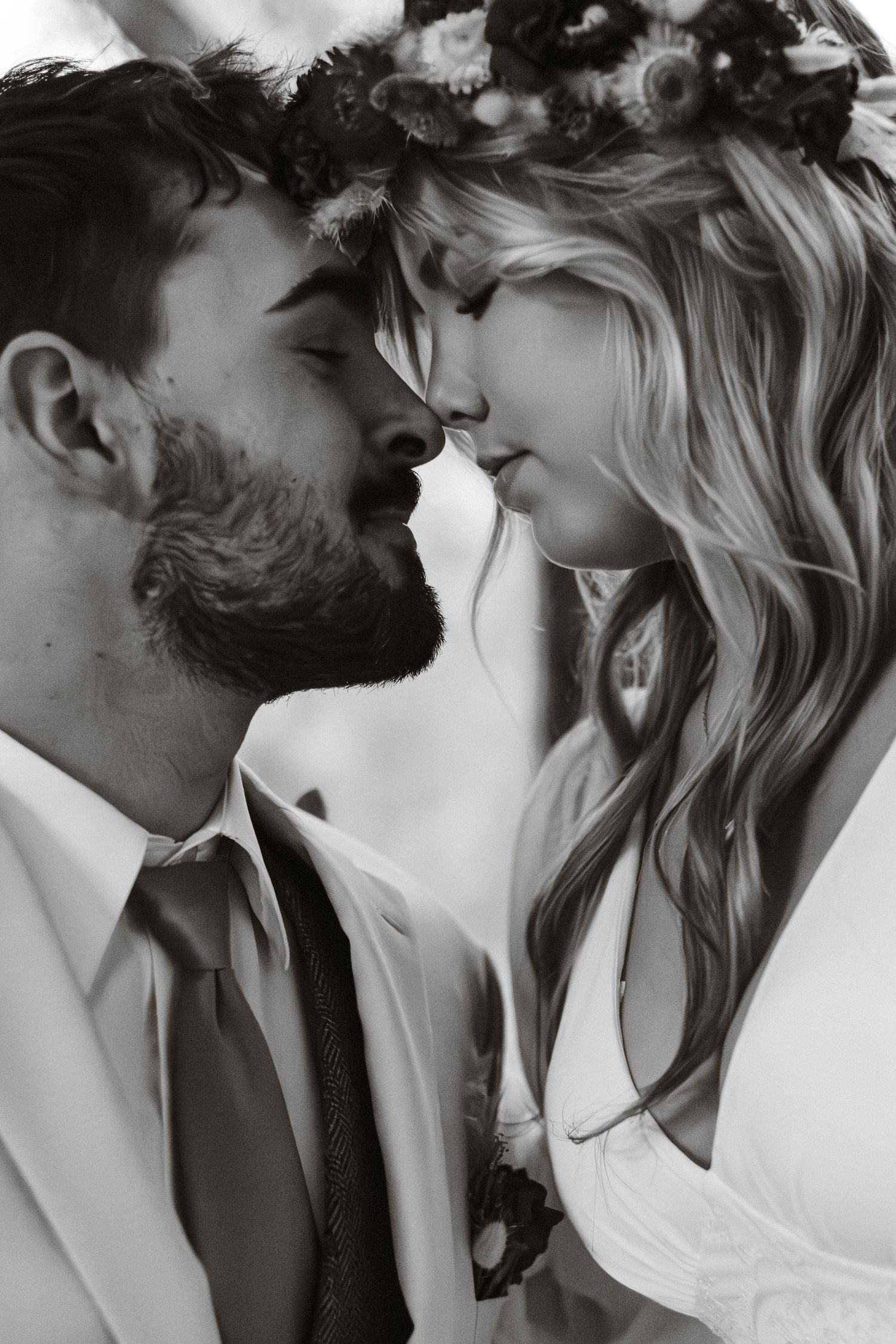 Close-up of bride and groom's faces nearly touching, she wears floral crown in black and white image.