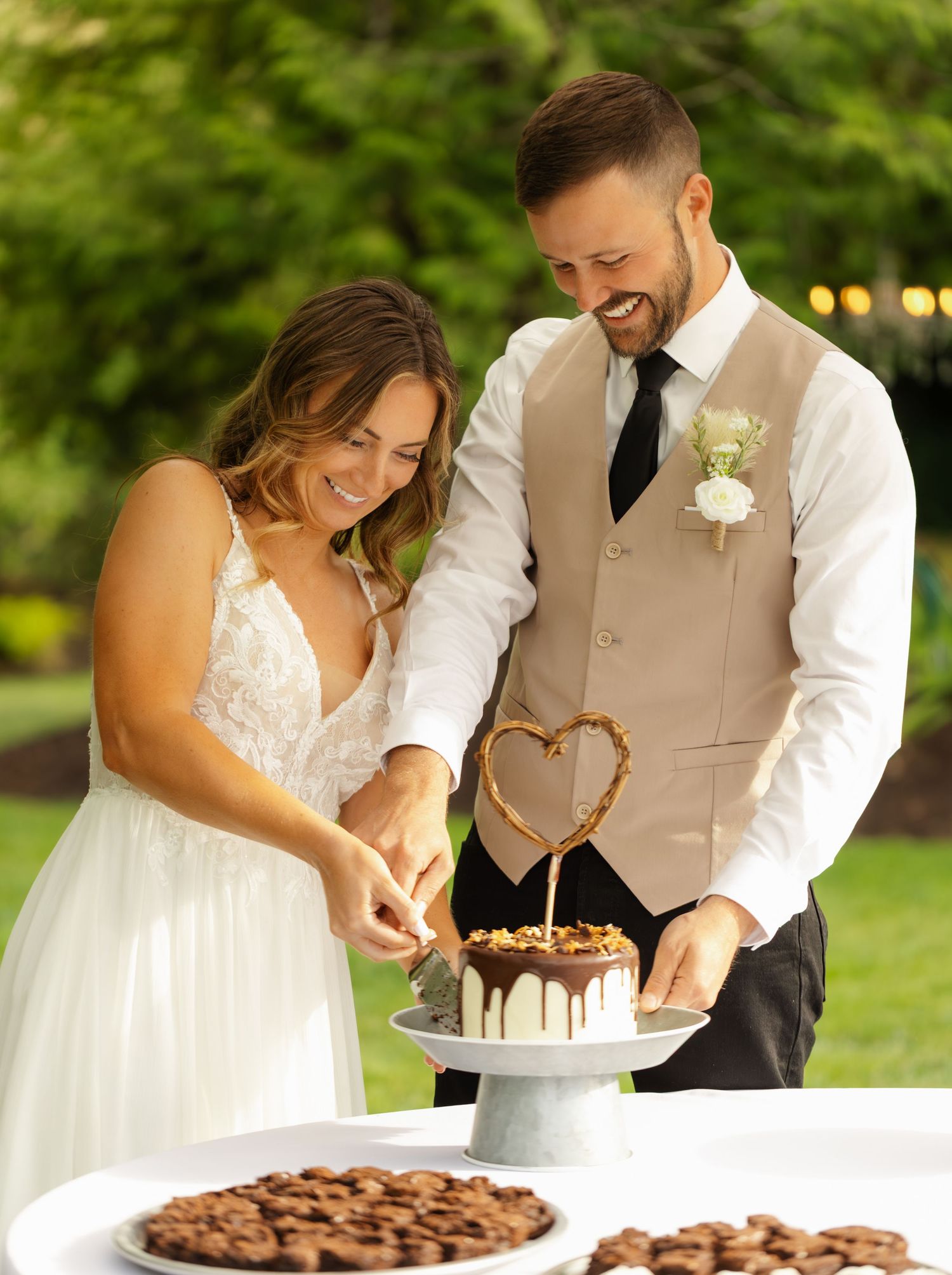 A bride and groom cut their rustic chocolate wedding cake together outdoors in a garden setting at sunset.
