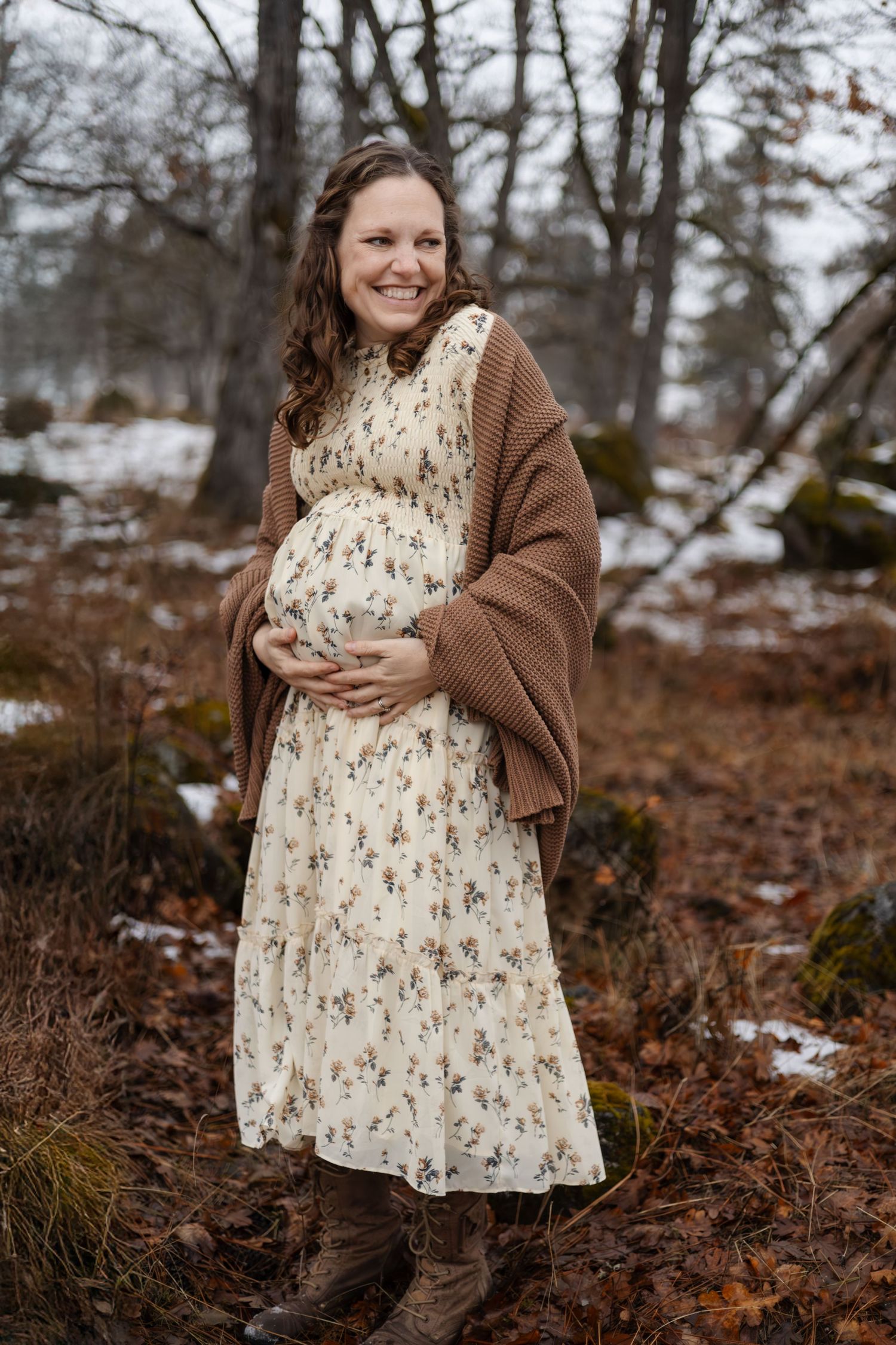 Winter maternity portrait in floral dress and brown cardigan among bare trees.
