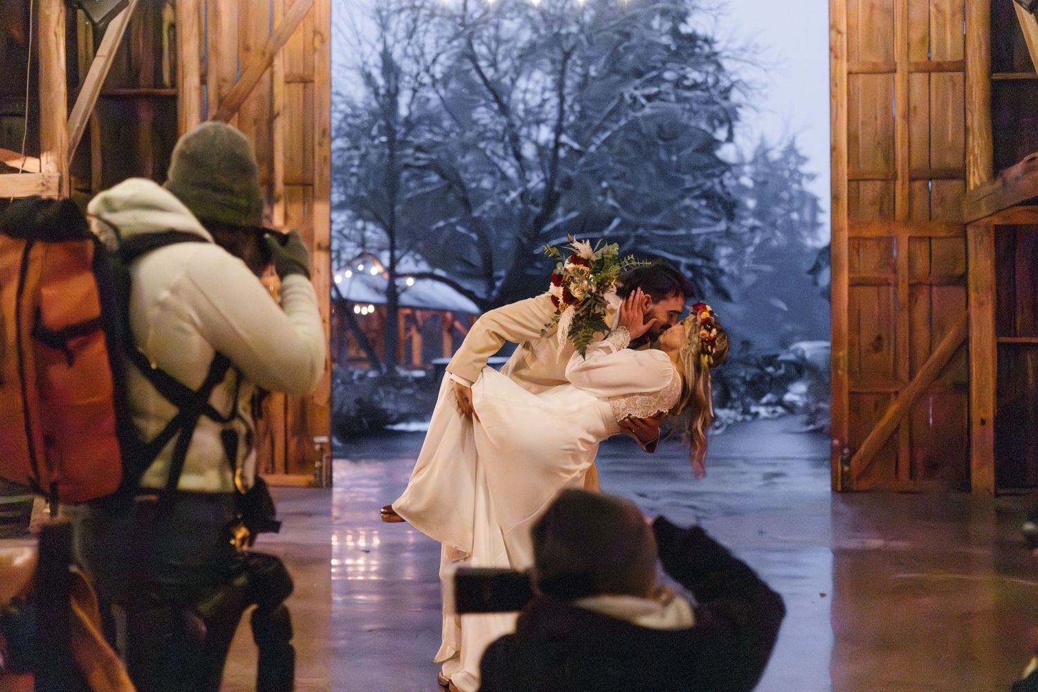 Groom dips bride for a kiss in rustic barn doorway, winter scene visible outside.