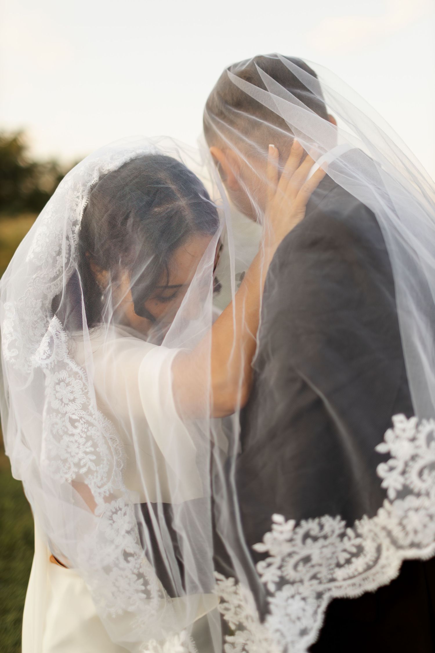 A romantic wedding moment captured as a couple shares an intimate kiss beneath a delicate lace veil at sunset.