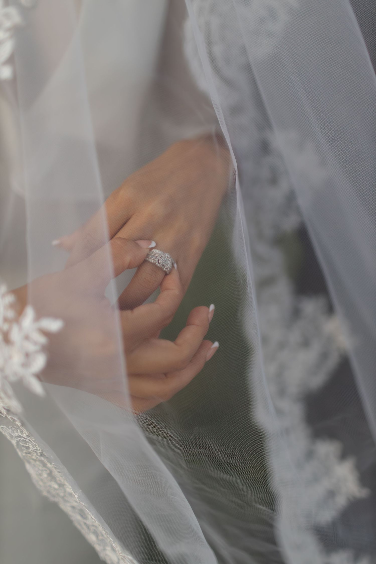 A delicate bridal veil frames an elegant engagement ring and manicured hand in soft lighting.