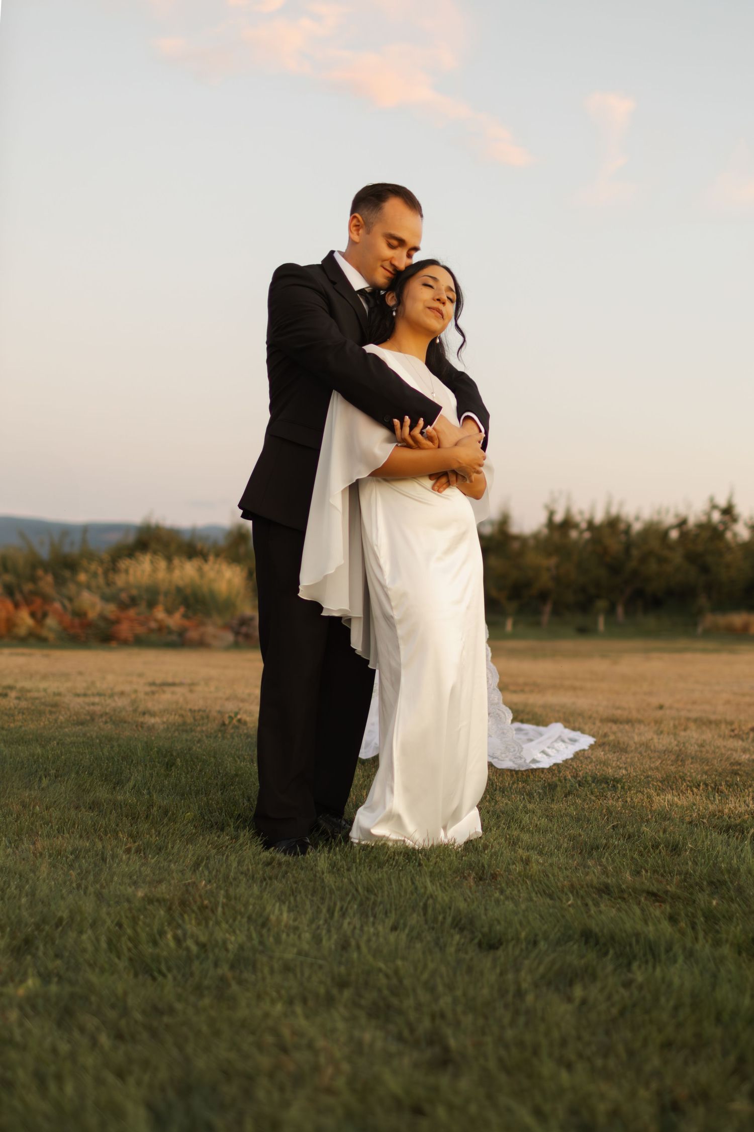 A couple in wedding attire embraces in a golden field at sunset, creating a romantic silhouette against the landscape.