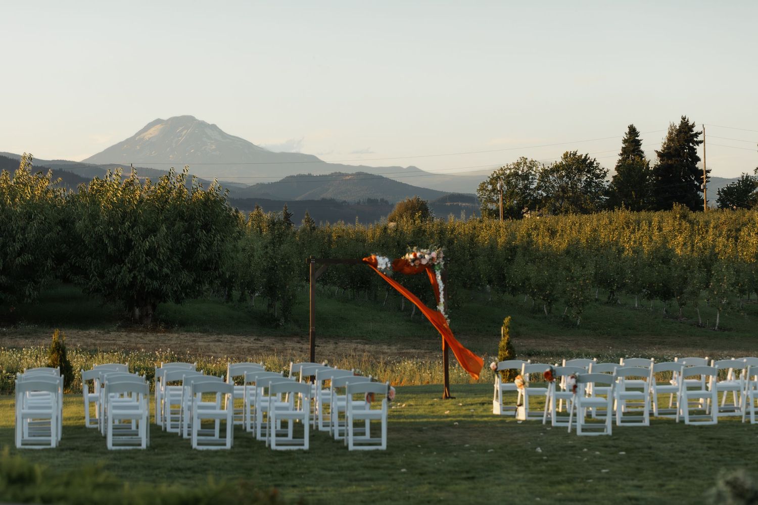 White wooden chairs arranged in rows on a grassy hillside with mountains and sunset in the background.
