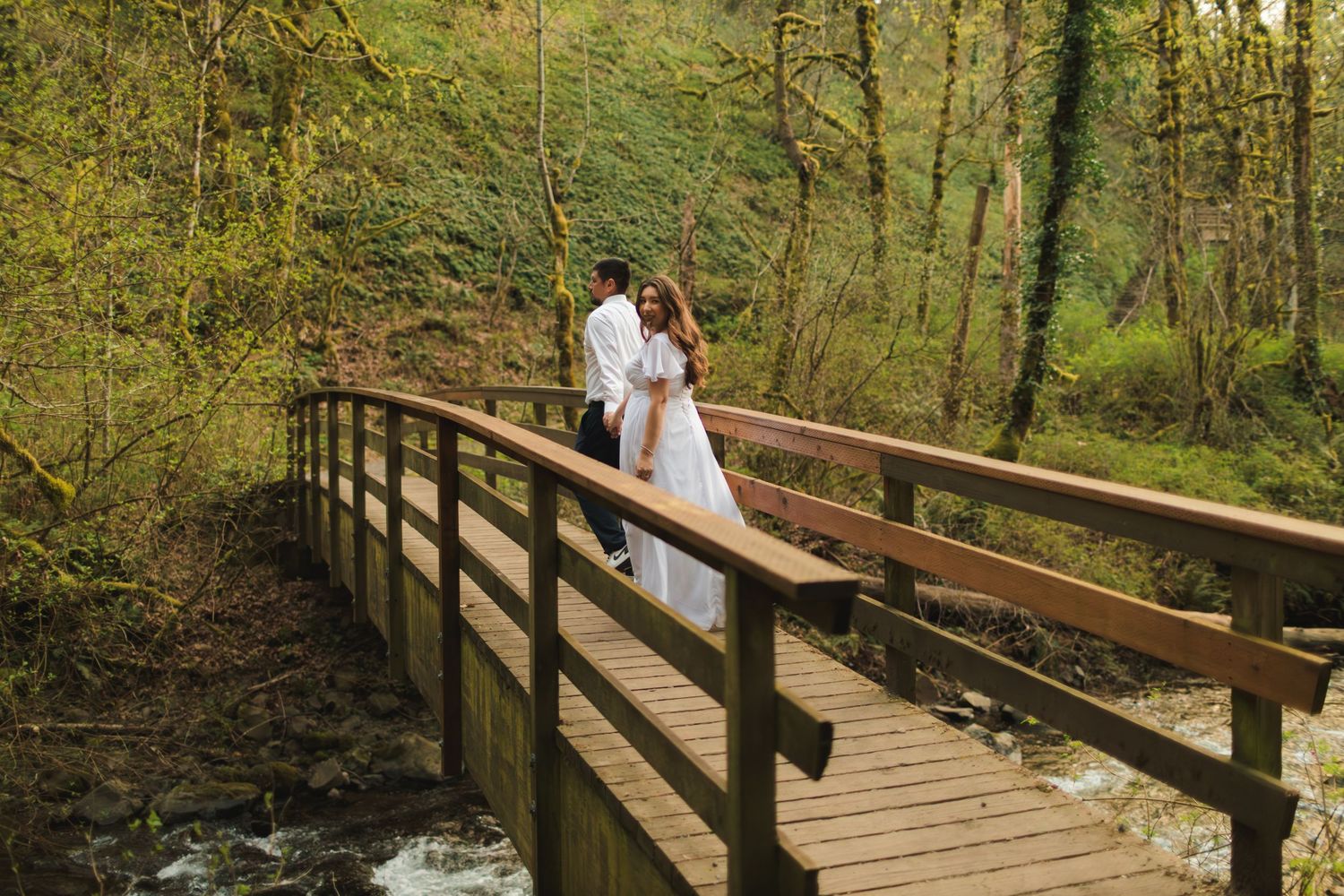 Wedding couple walking on a wooden bridge over a stream surrounded by lush green trees.