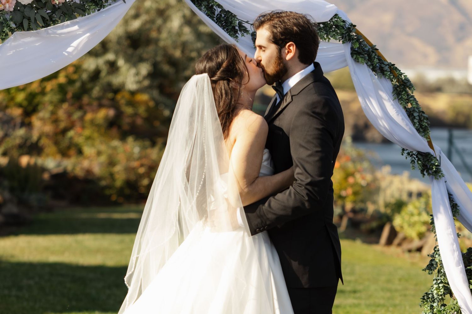 Bride and groom share first kiss at outdoor wedding ceremony beneath floral arch with mountain views.