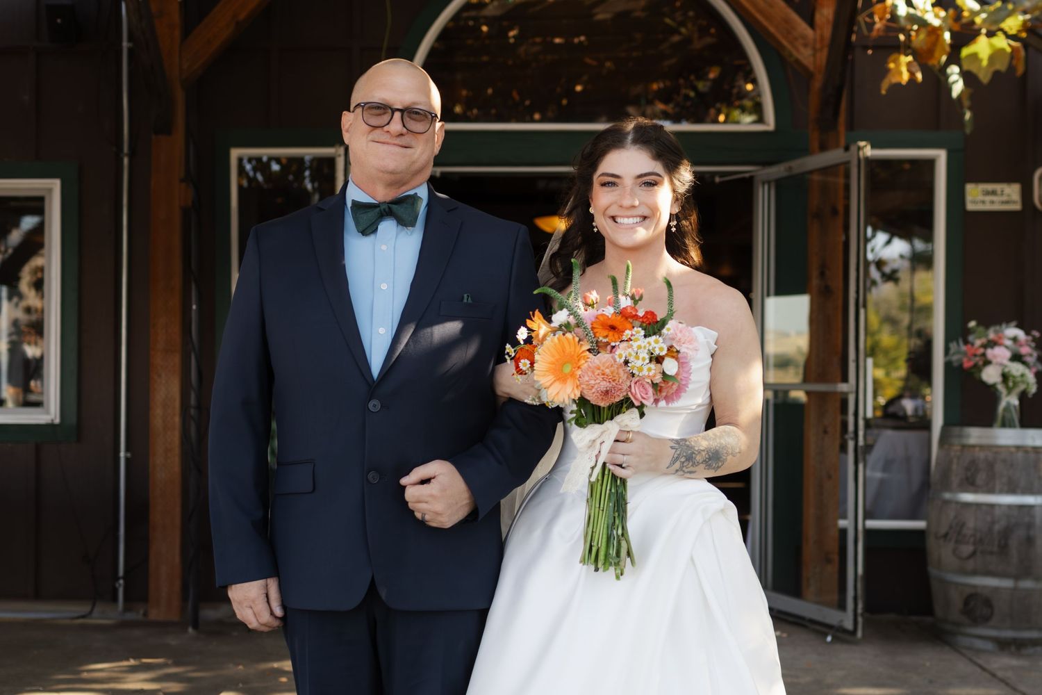 Bride in white gown holding colorful bouquet stands with suited man in bow tie outside rustic wedding venue.
