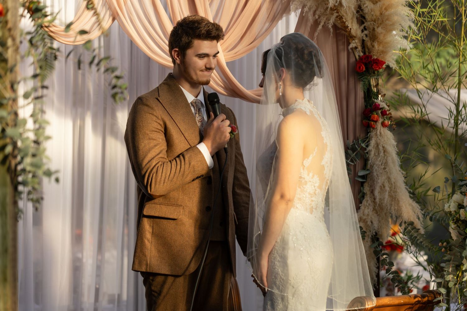 A groom in a brown suit holds a microphone while reciting vows to a bride in a white lace gown and veil.