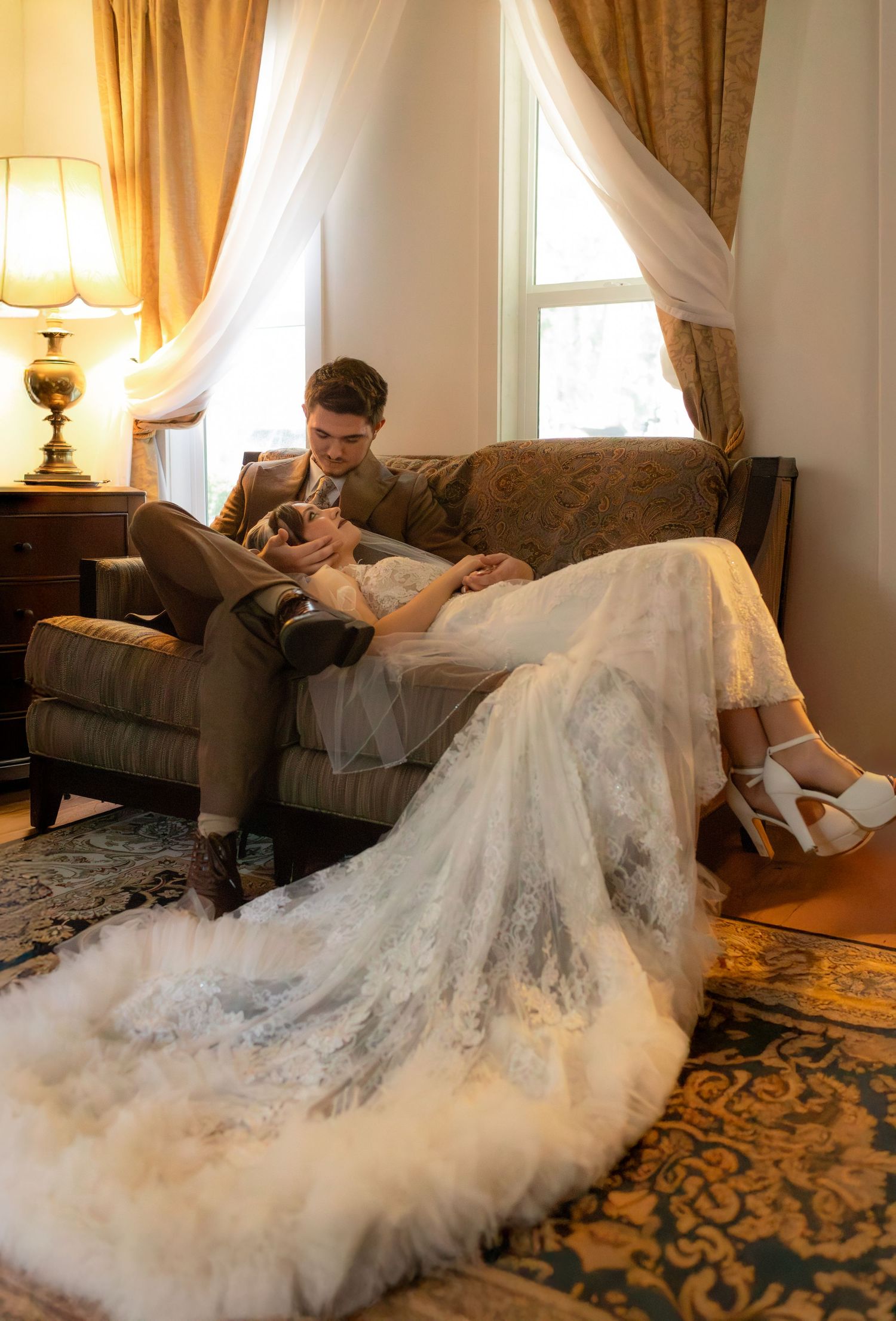 A romantic wedding photo of a couple lounging on an elegant couch with a flowing white lace dress trailing on the floor.