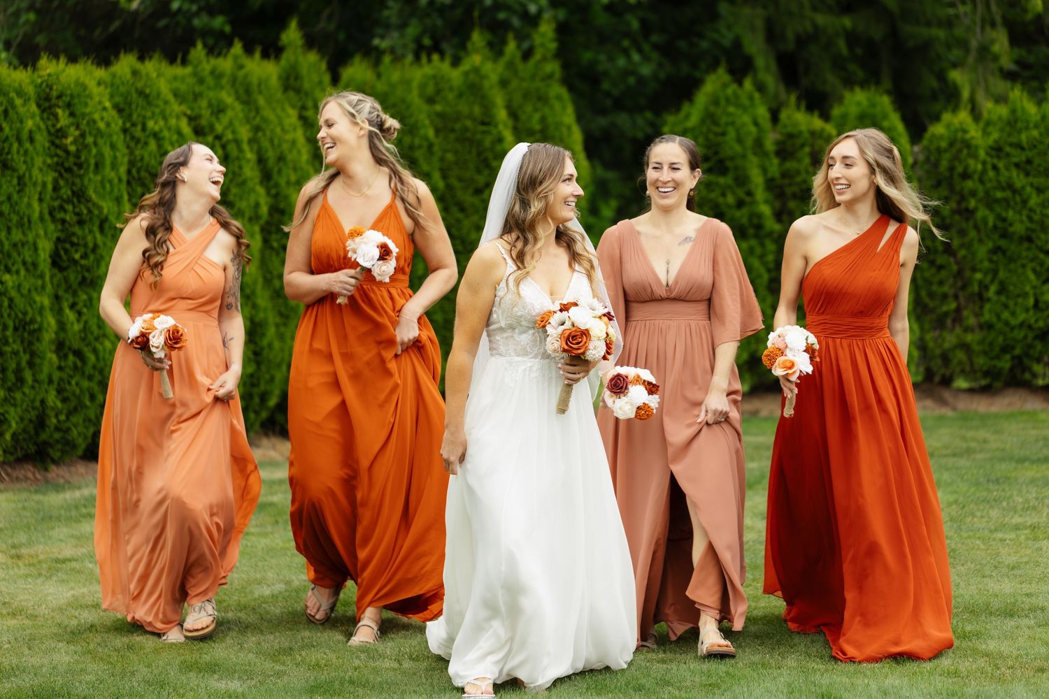 A wedding party in white and orange dresses walks together on grass during an outdoor celebration.