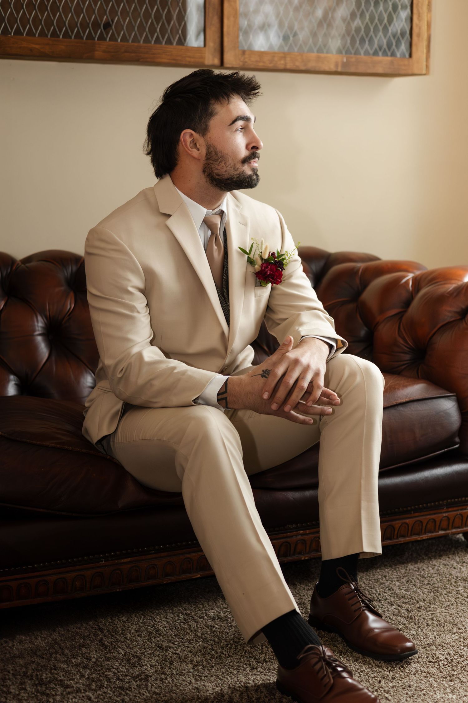 Groom in tan suit sits on leather couch, looking pensively to the side with a boutonniere on lapel.