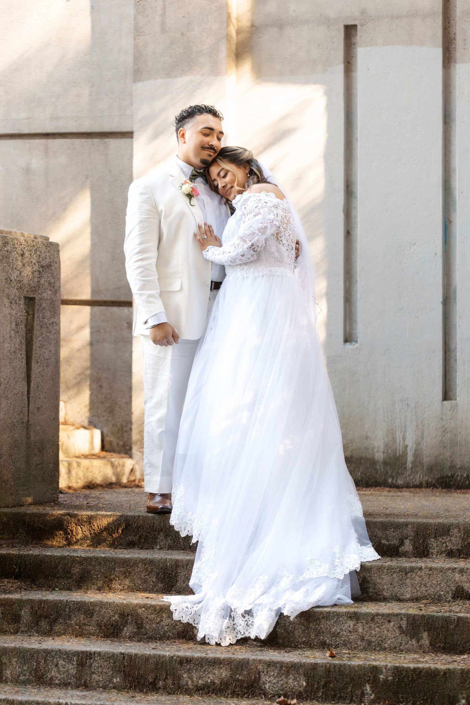 A couple in wedding attire poses on concrete steps during golden hour.