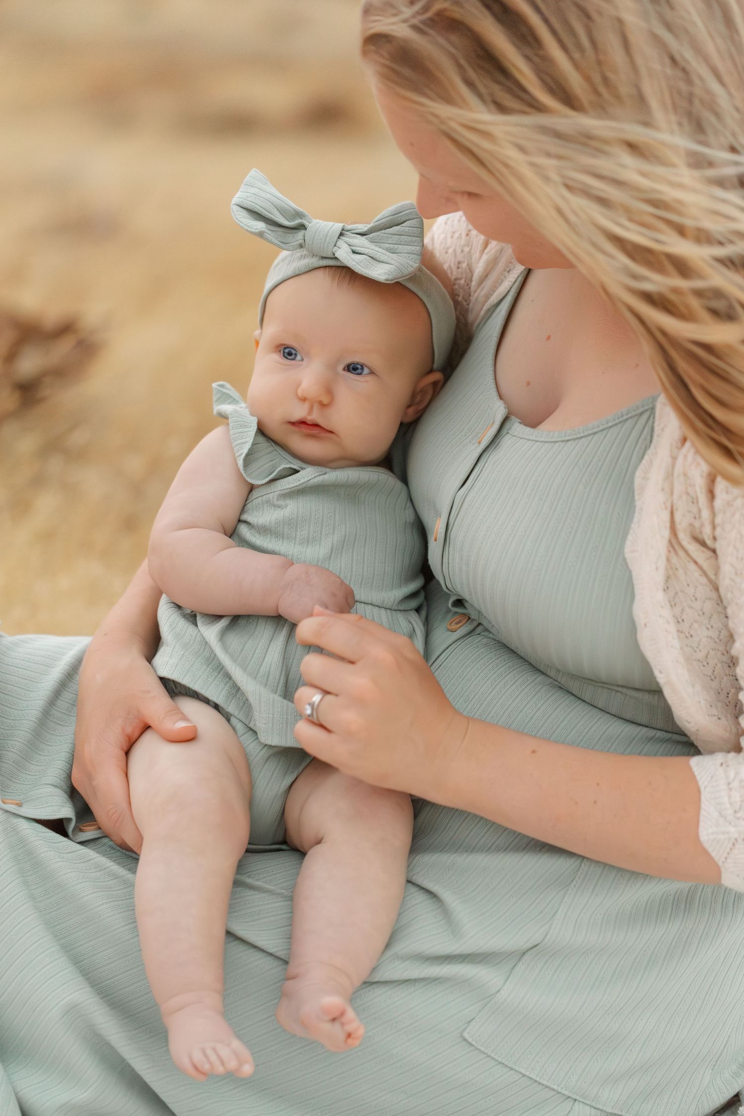 Baby in sage green romper being held on mother's lap in soft natural lighting.