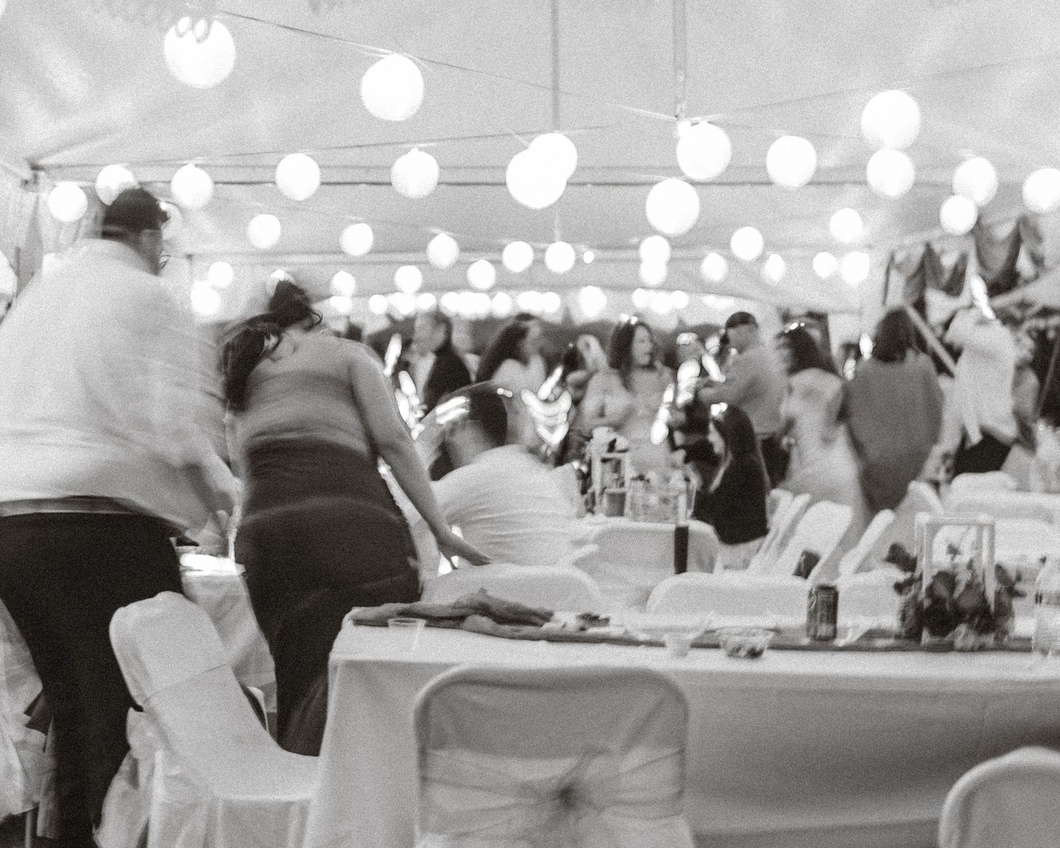 A black and white panoramic view of a wedding reception with guests seated at tables under string lights.