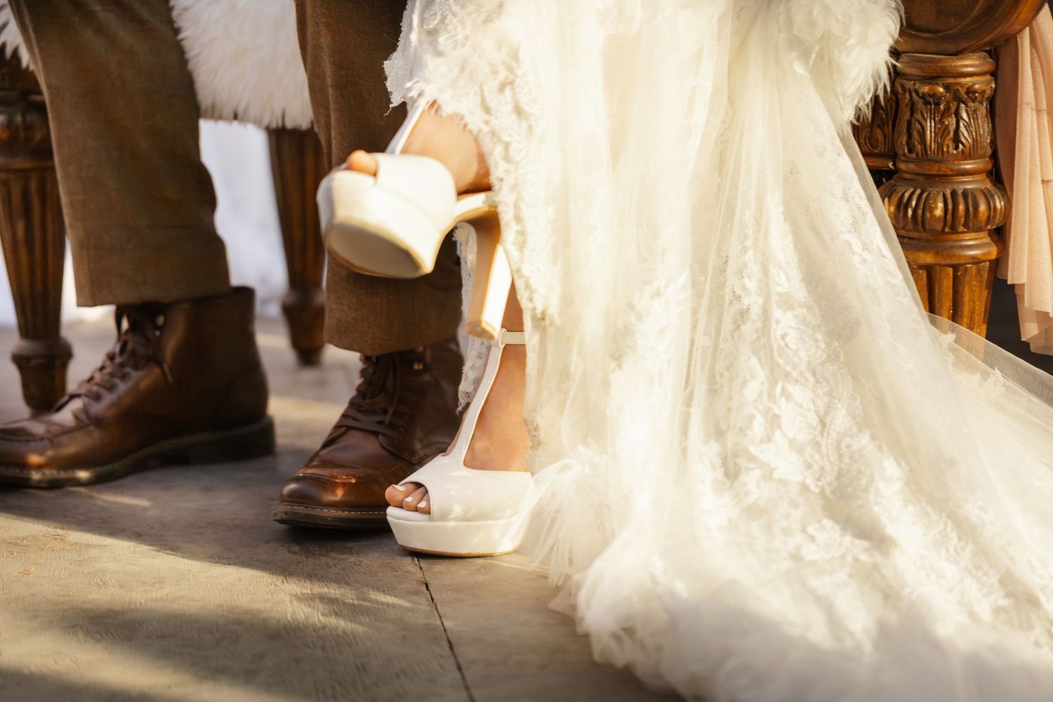 Close-up view of brown leather boots and flowing white wedding dress hem on wooden floor.