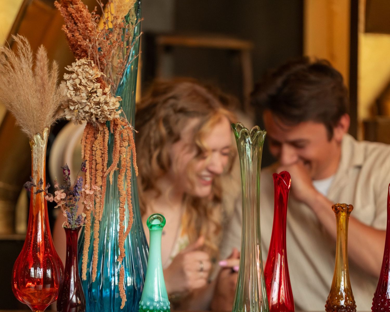 Couple smiling behind colorful glass bud vases and dried flowers in a warm, cozy shop setting.