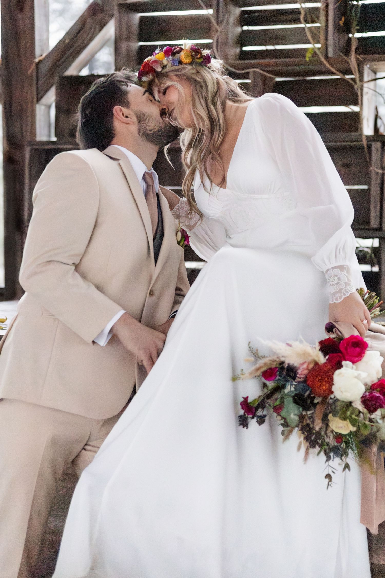 Bride and groom share tender kiss on wooden steps, she in white dress with floral crown, he in light suit.