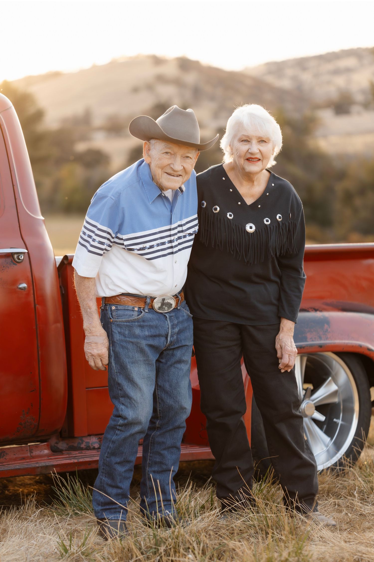 A senior couple stands together by a classic red truck in a golden field.