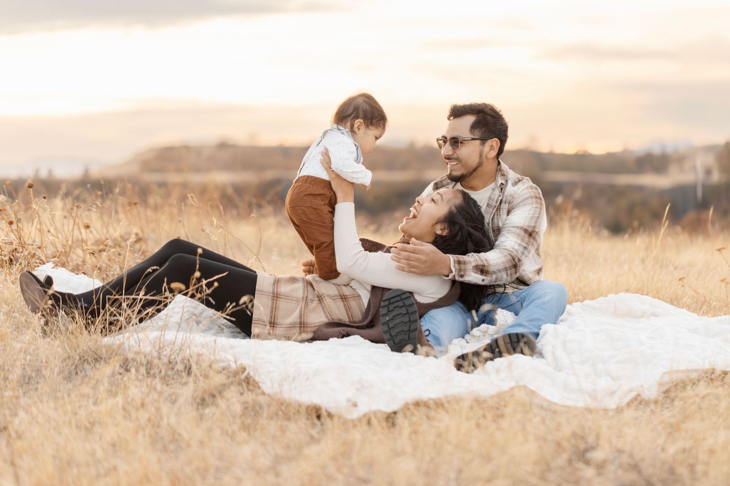 Family enjoying a picnic together on white blanket in golden field at sunset.