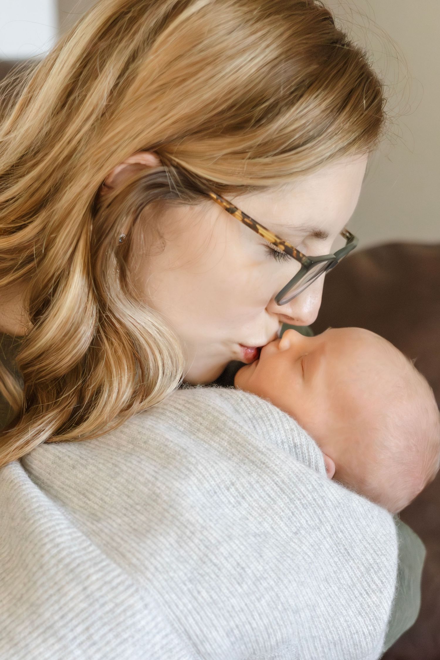 Woman wearing glasses gently embraces sleeping infant wrapped in light gray blanket.