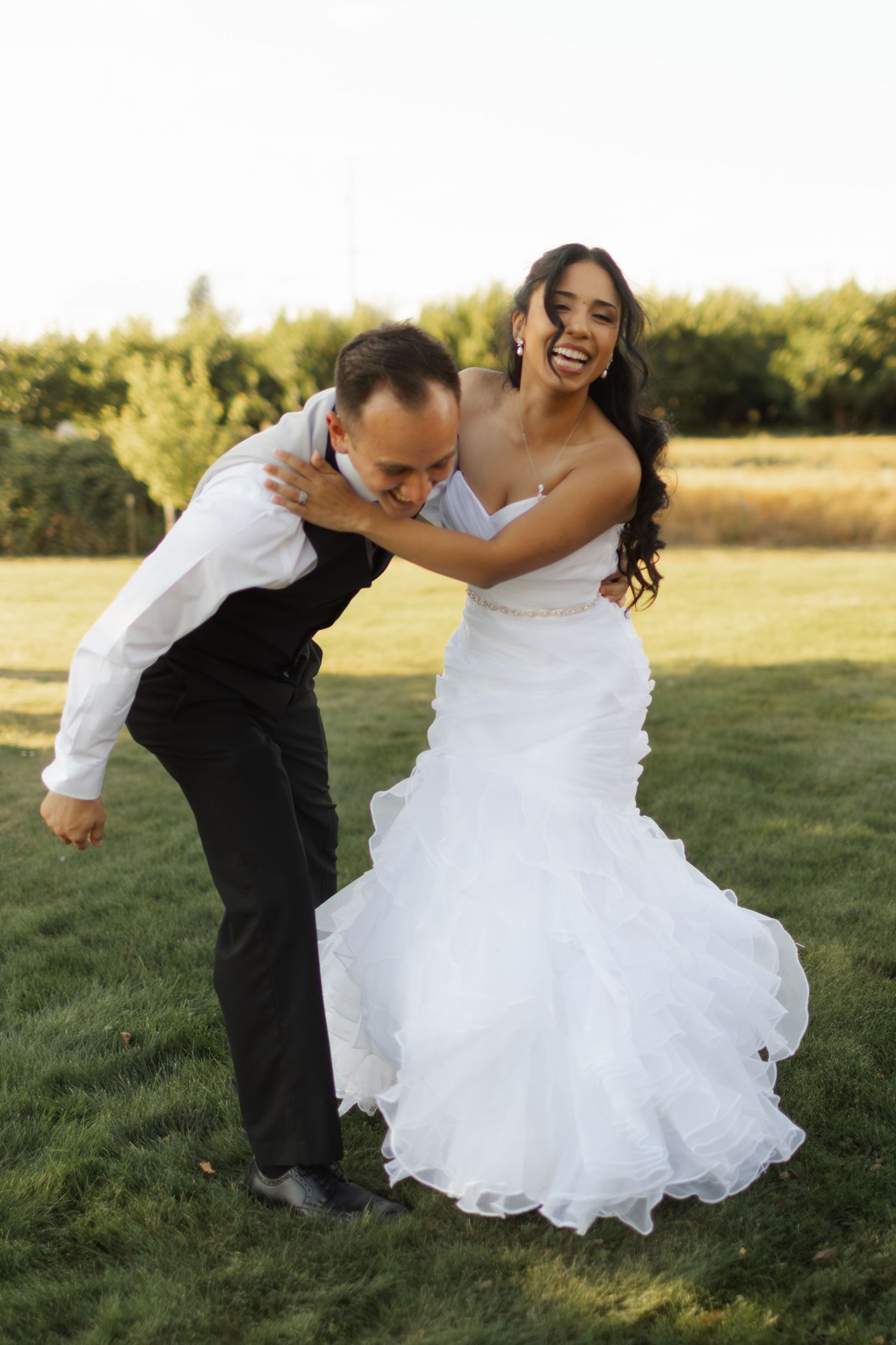 A couple in wedding attire playfully dips and dances together in a sunny outdoor field at sunset.
