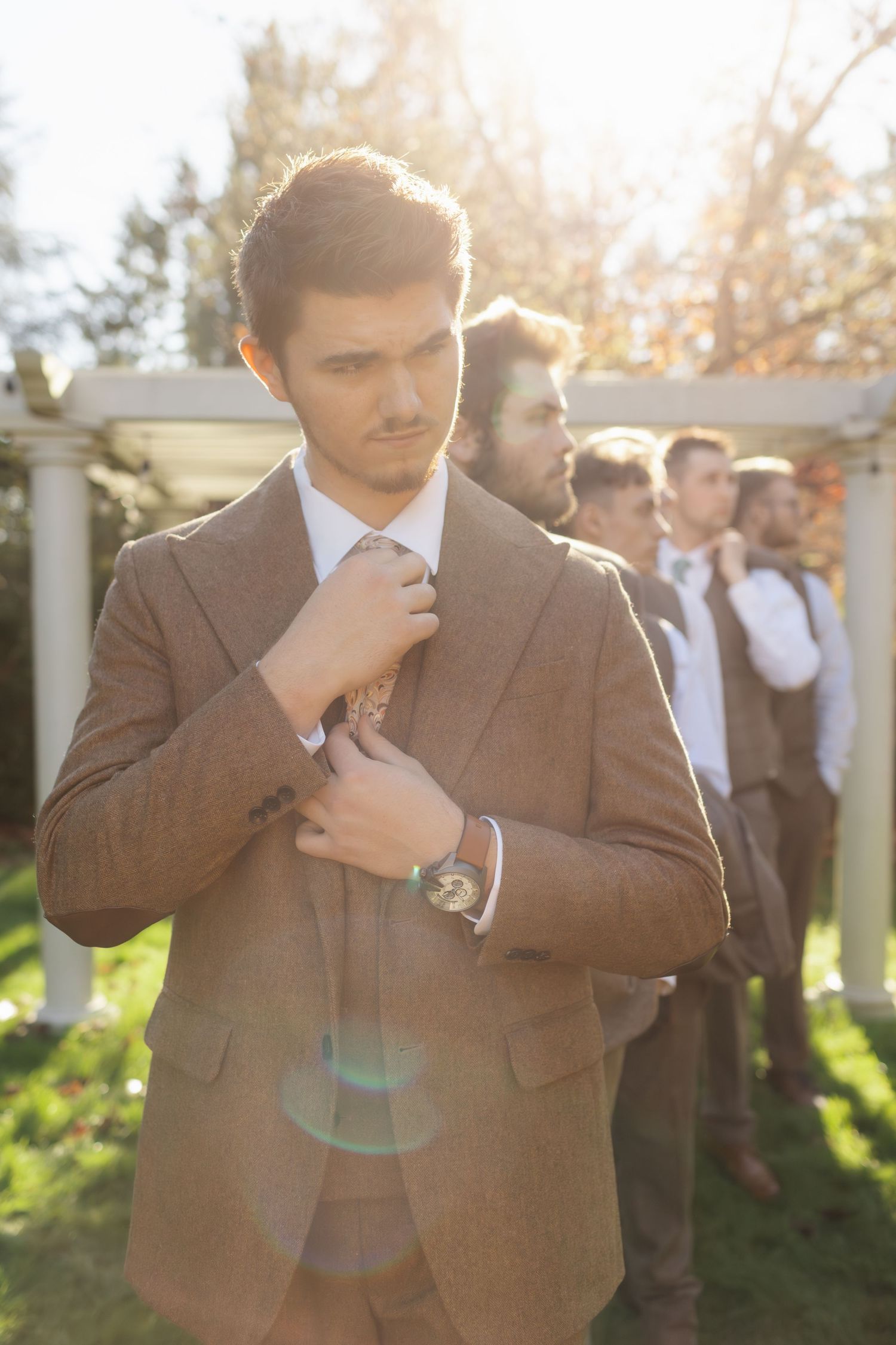 A person in a brown suit adjusts their tie while standing outdoors in warm sunlight with others visible in background.