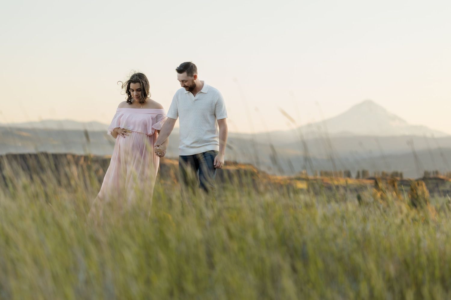 Romantic couple embraces in a golden field at sunset with mountains in the background.
