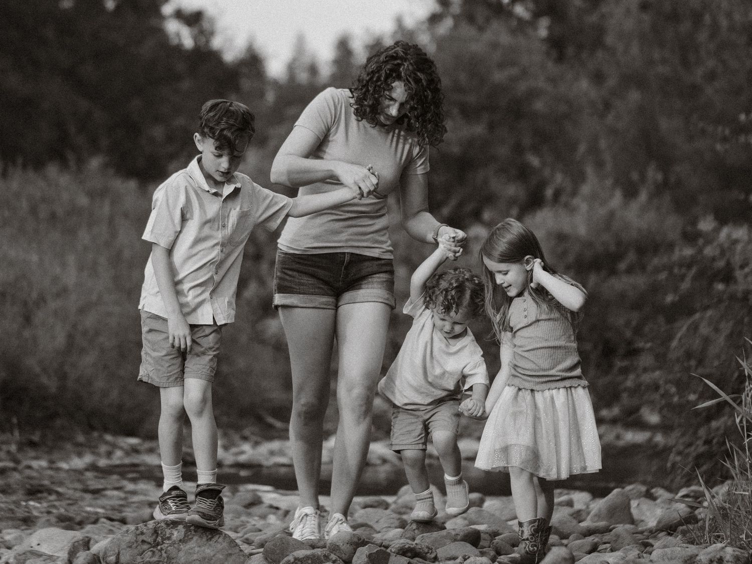 Black and white photo of a woman helping three young children walk across rocky terrain outdoors.