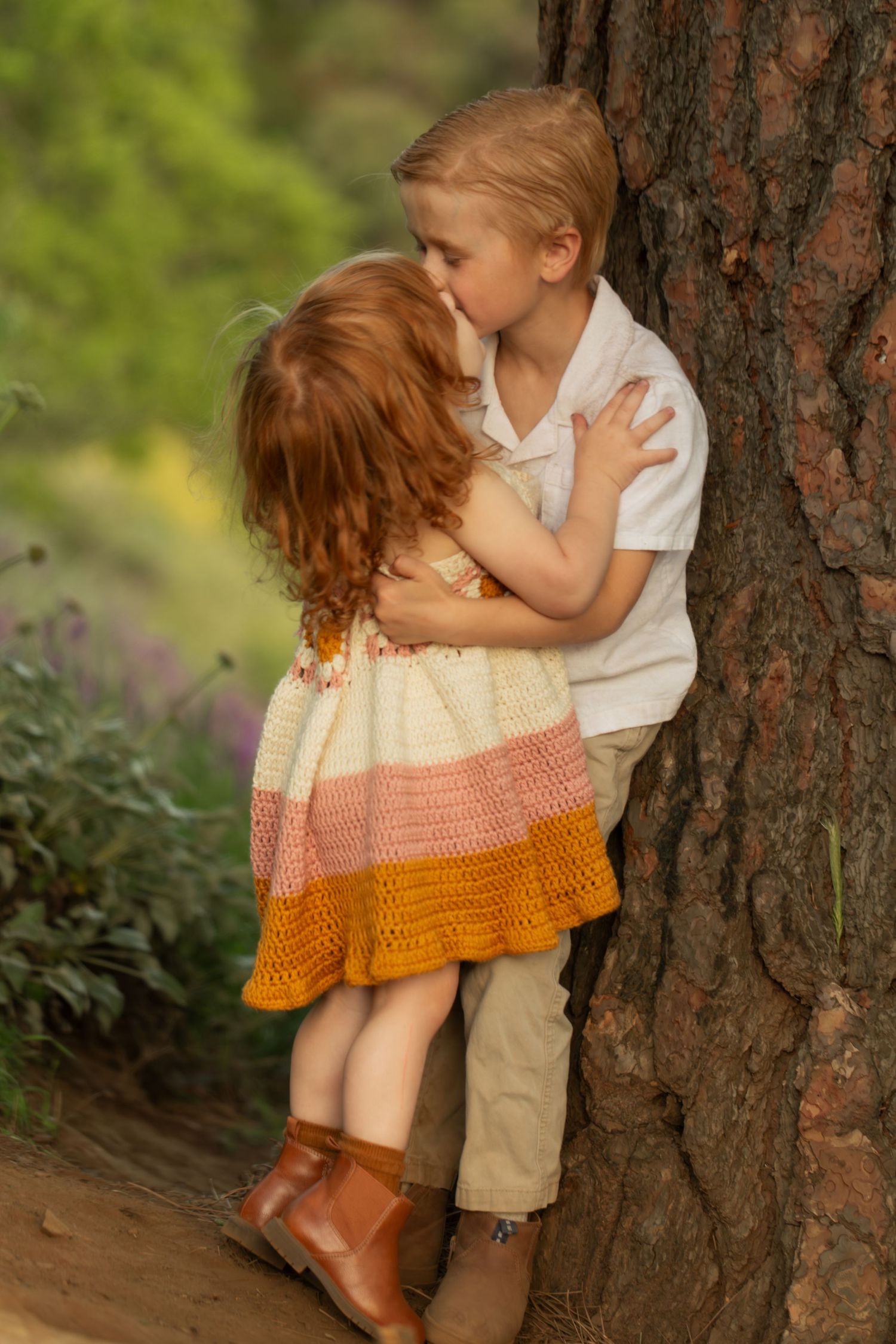 Two young children share a sweet hug near a tree in a lush garden, girl in colorful crochet dress, boy in white shirt.