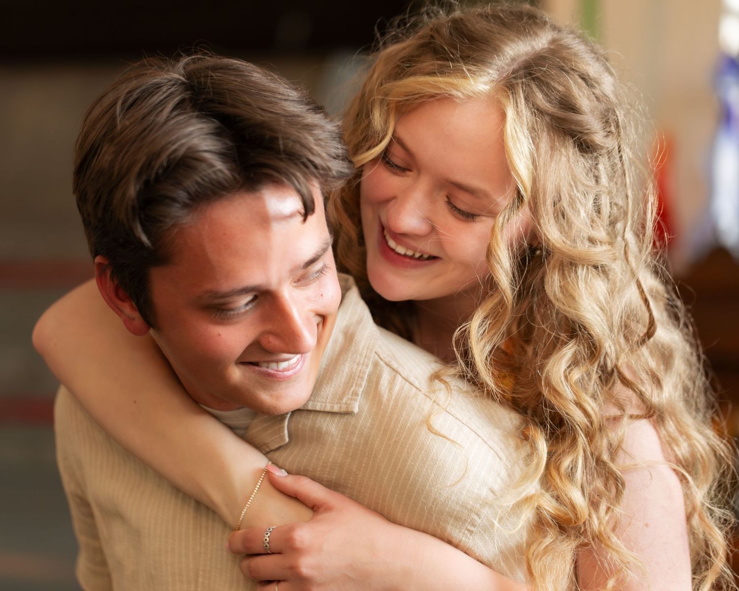 Young couple smiling and embracing warmly, woman with curly blonde hair hugging man from behind in cozy indoor setting.