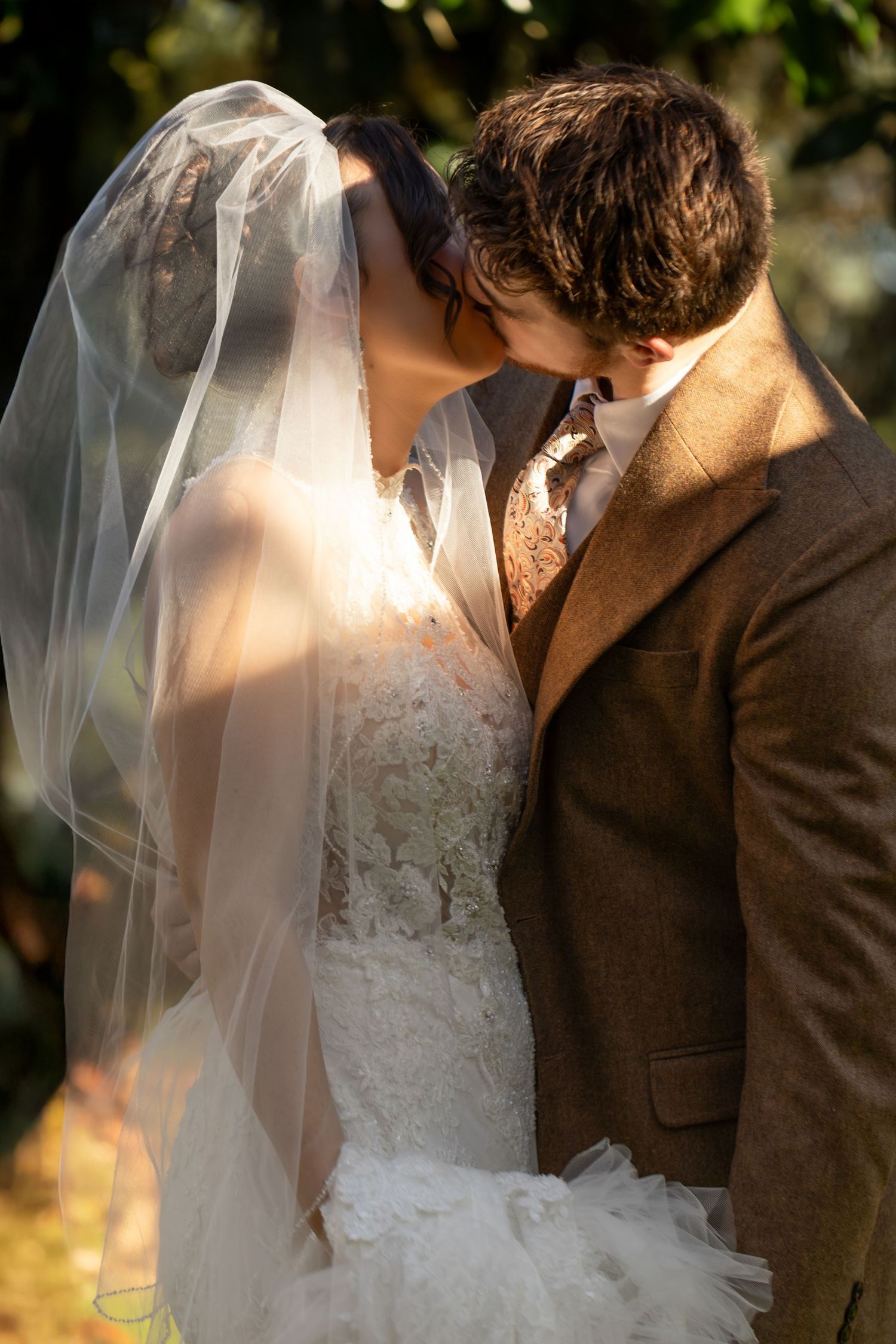 Romantic wedding couple embraces in soft natural lighting with bride wearing white lace gown and flowing veil.