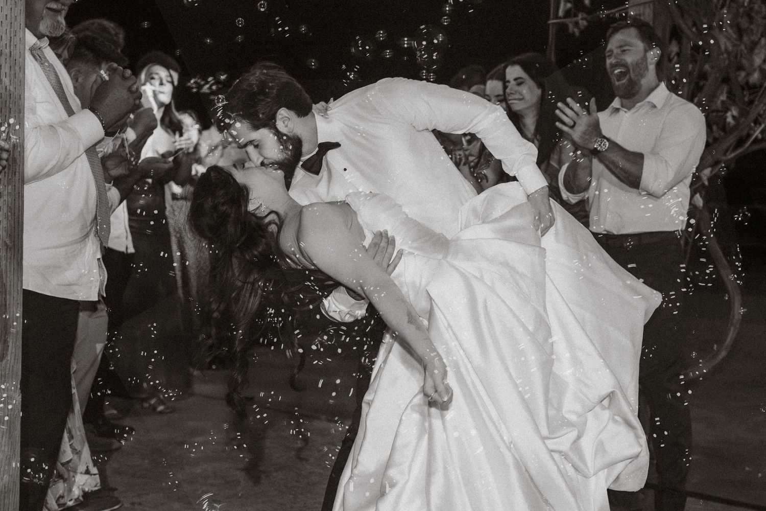 Wedding guests celebrate with bubbles as newlyweds dance together in black and white photography.