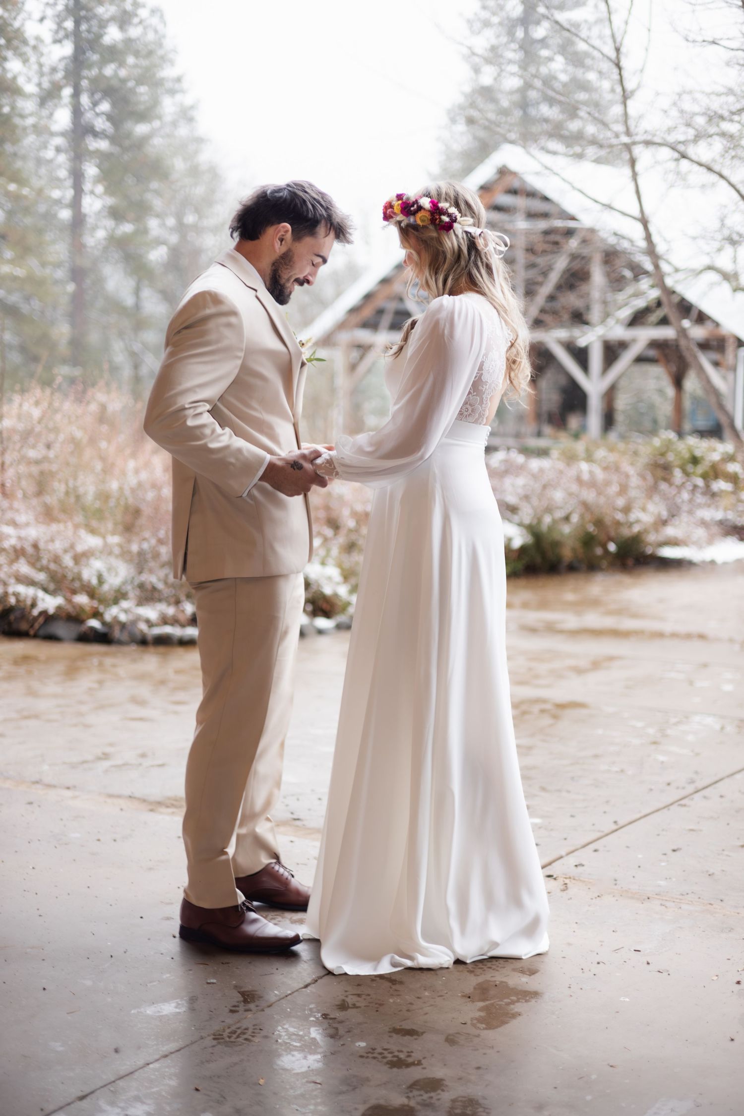 Bride and groom hold hands outdoors, she in white dress with floral crown, he in tan suit against snowy backdrop.