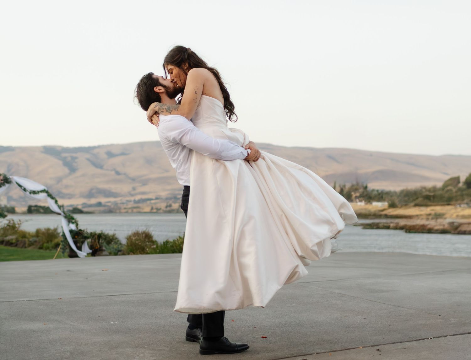 Groom lifting bride in white wedding dress, sharing romantic kiss outdoors with scenic hills and lake backdrop.