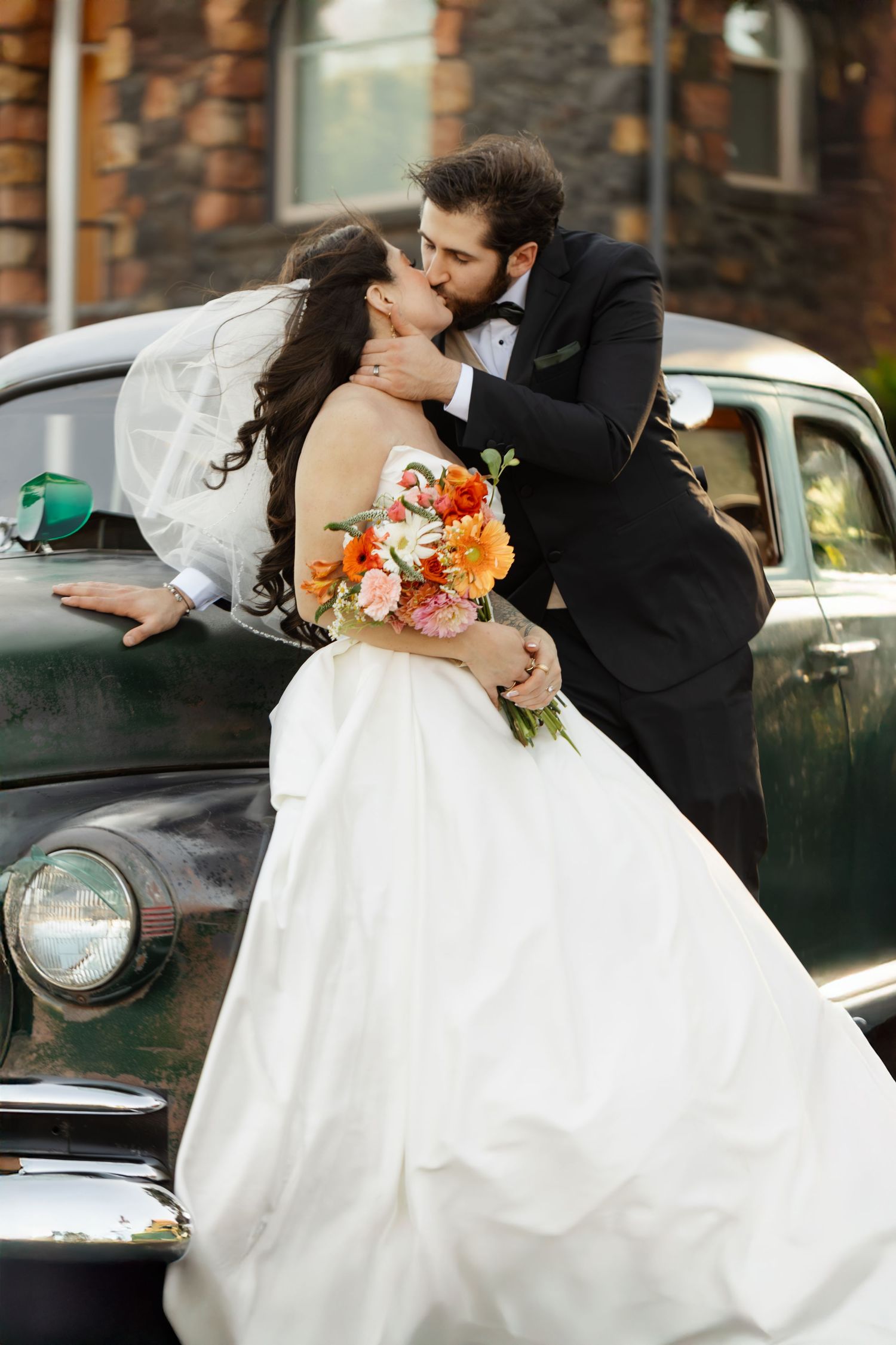 Romantic wedding couple embraces in front of vintage cars during their outdoor photo session.