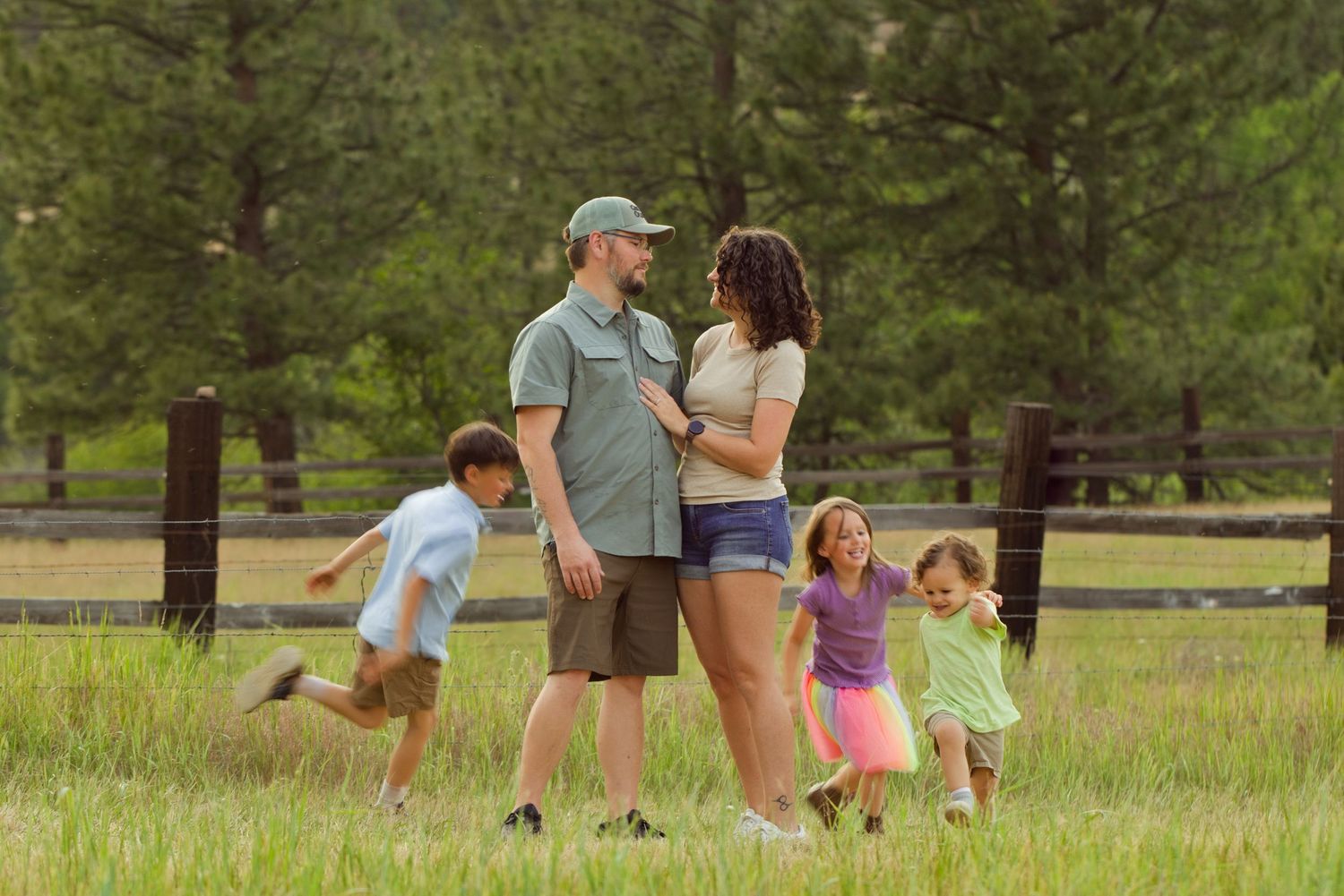 Happy family with two young children playing in a green meadow with pine trees and wooden fence.