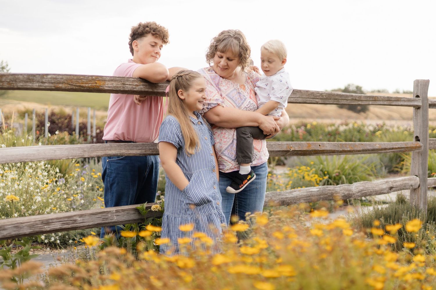 Family members share tender moments among blooming wildflowers near wooden fence.