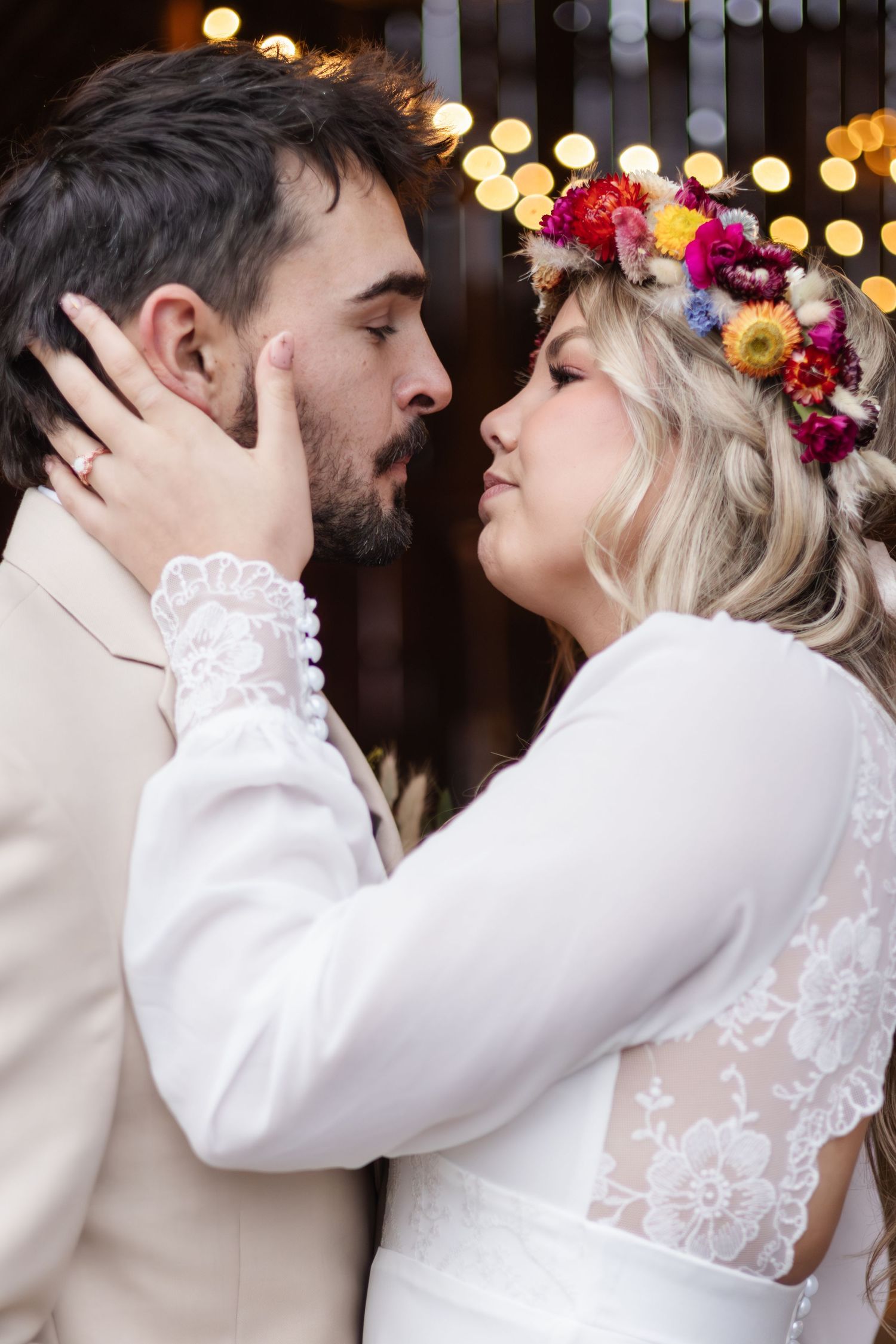 Bride and groom in romantic embrace, woman wearing floral crown and lace dress against twinkling lights background.