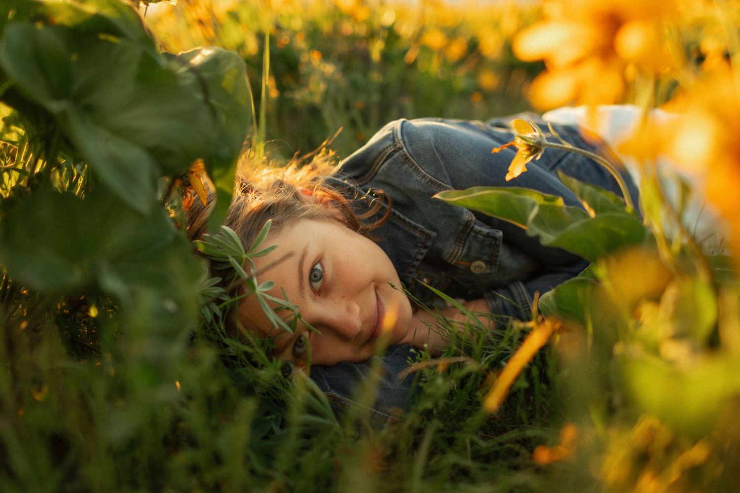 A young woman lying in a lush green field surrounded by golden yellow flowers in warm sunlight.
