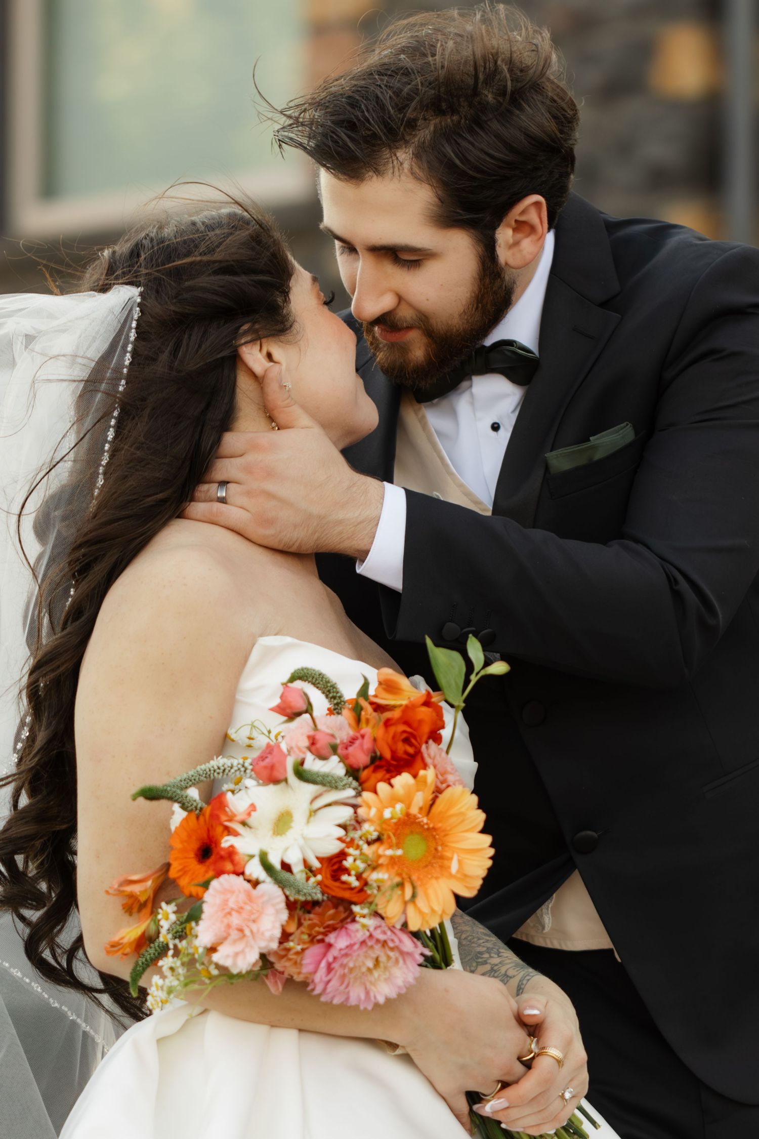Romantic wedding couple sharing intimate kisses while bride holds vibrant orange and pink floral bouquet.