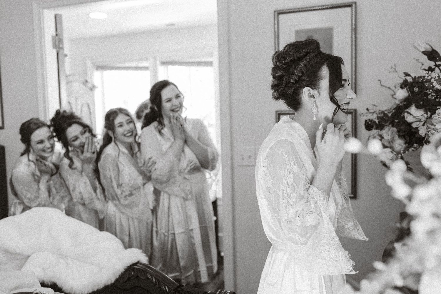 Black and white photo of a bride in a lace robe crying while bridesmaids in robes react emotionally behind her.