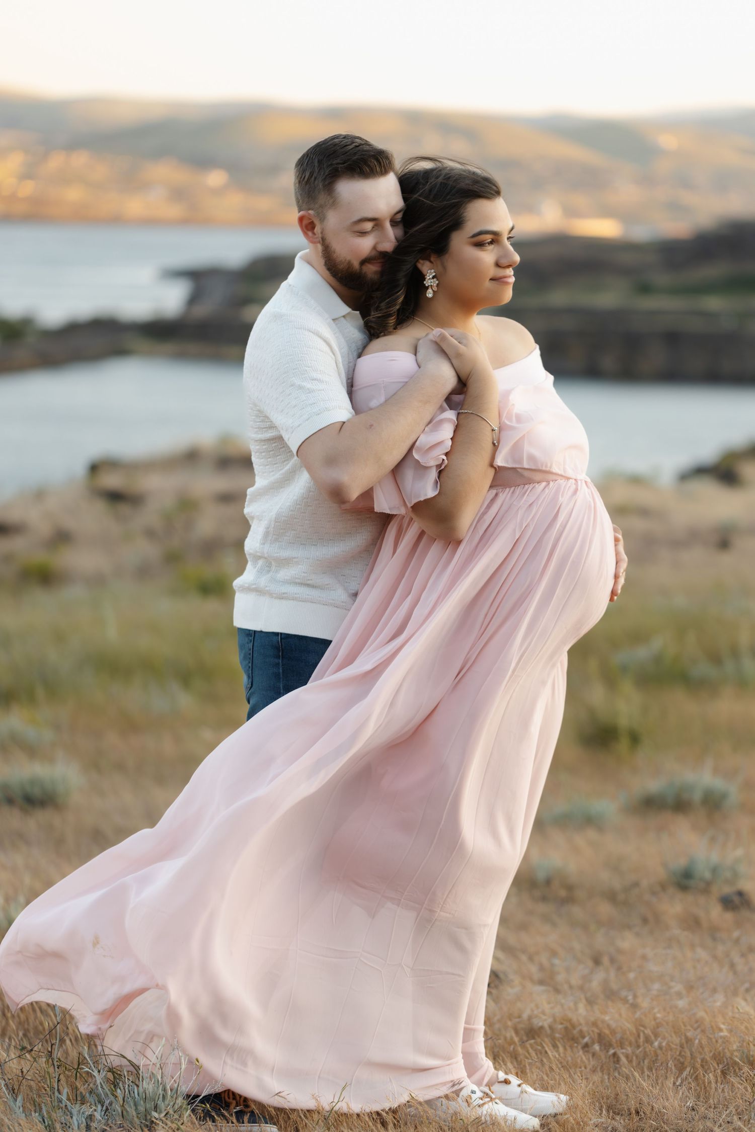 A couple embraces in a pink flowy dress against a scenic mountain landscape at sunset.
