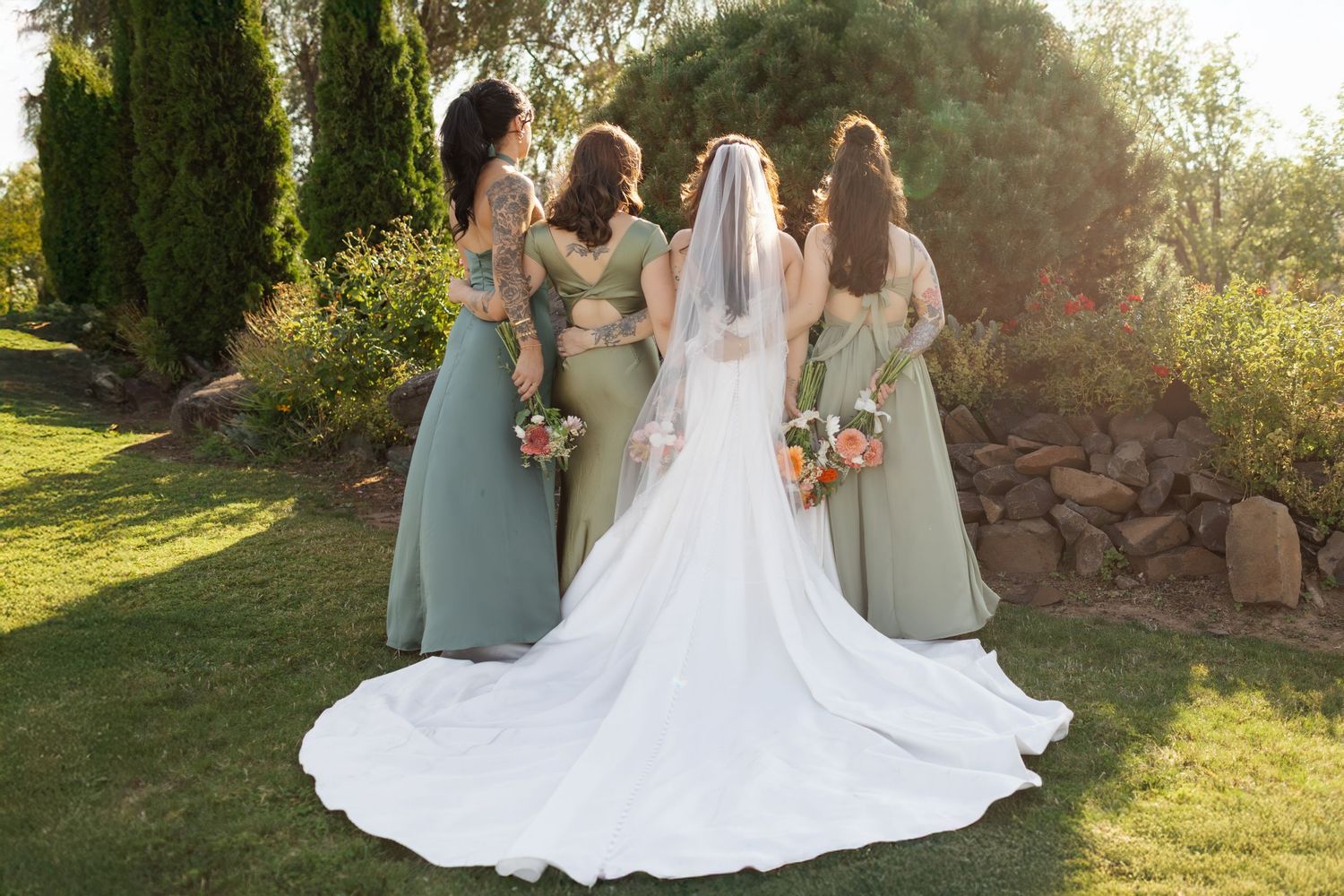 Bride in white gown with veil stands with three bridesmaids in sage green dresses holding bouquets in a garden.