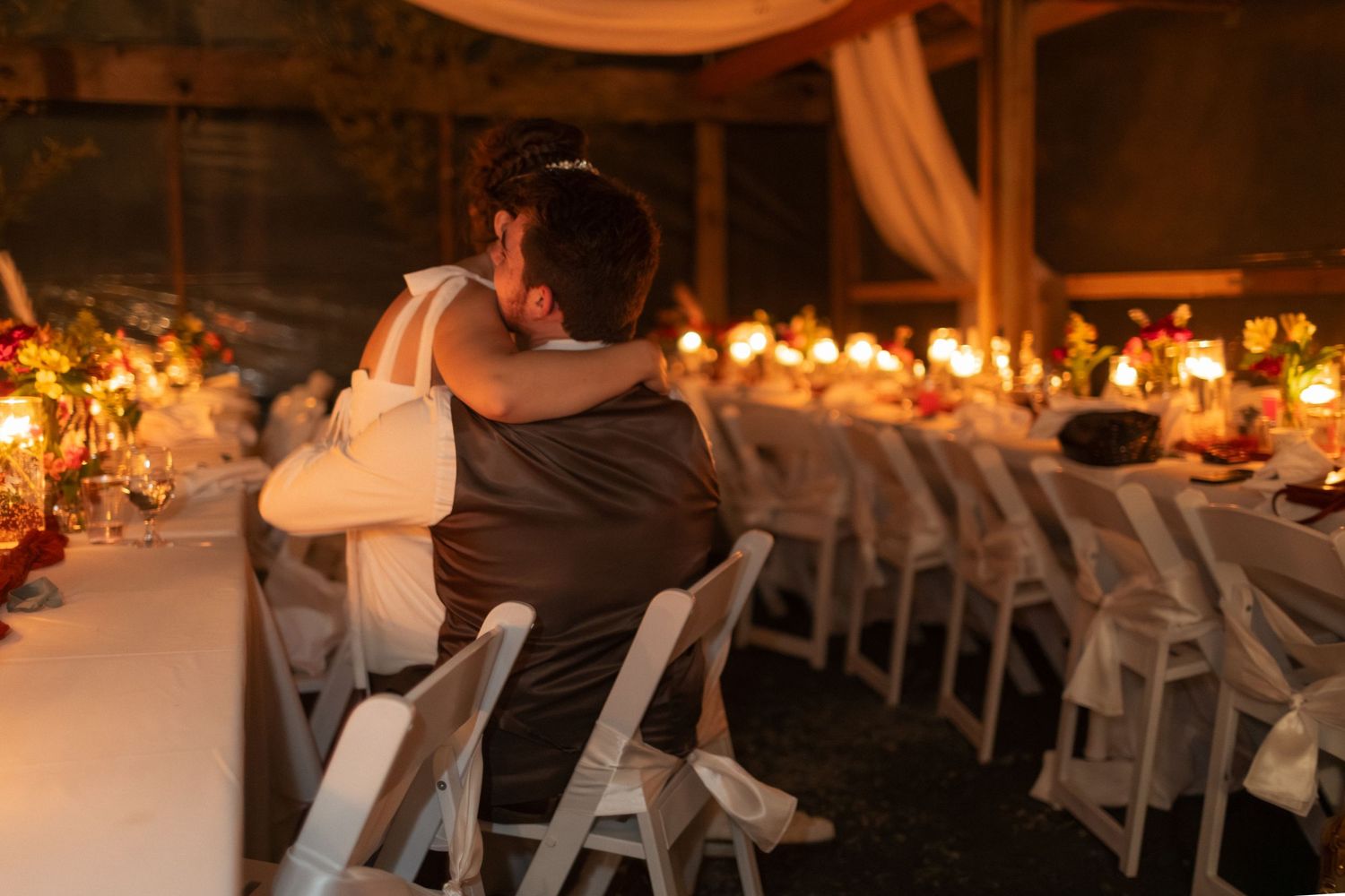 Couple embracing at candlelit wedding reception dinner table in warmly decorated venue.