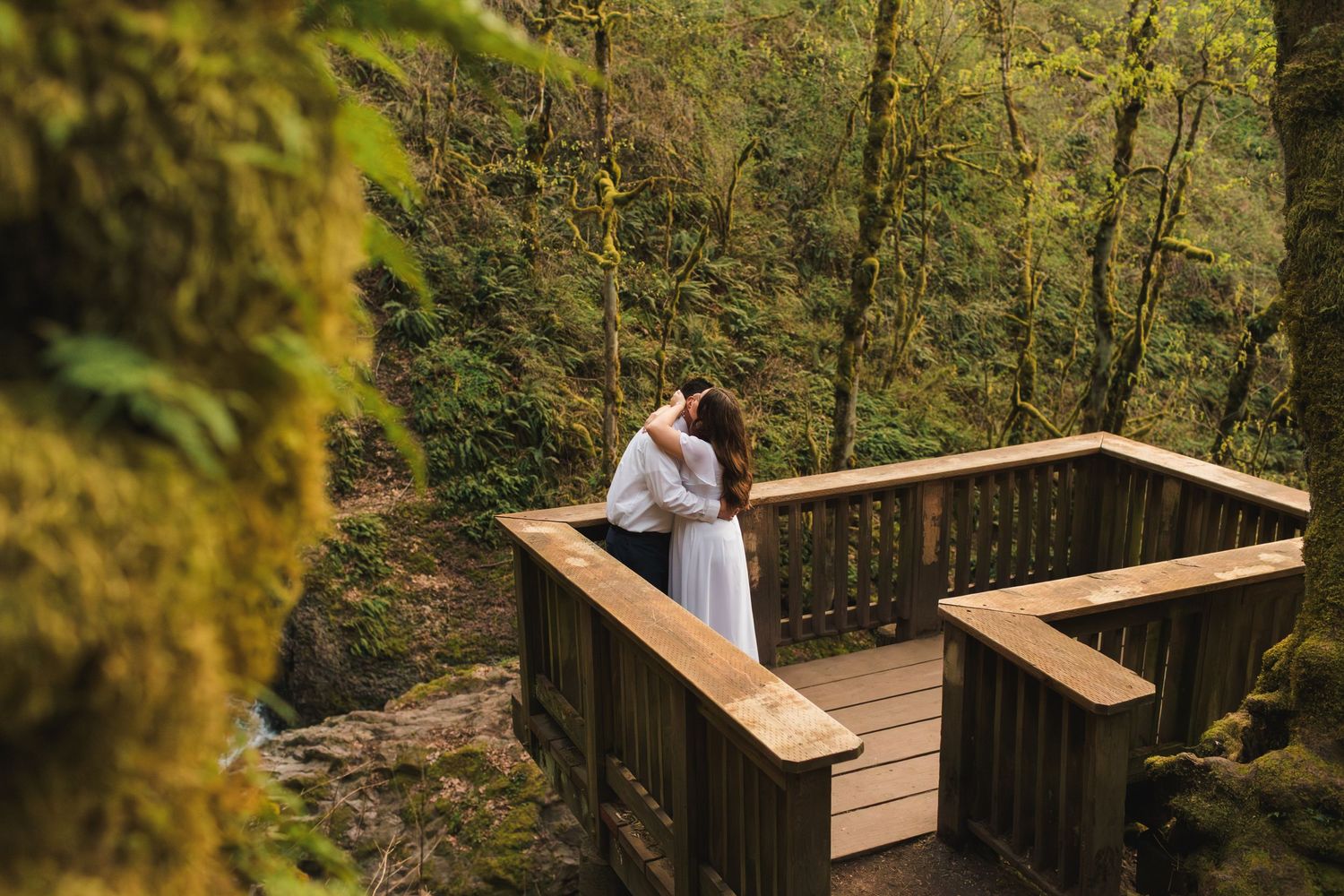Couple embracing on a wooden overlook deck surrounded by lush green mossy forest trees in a romantic outdoor setting.