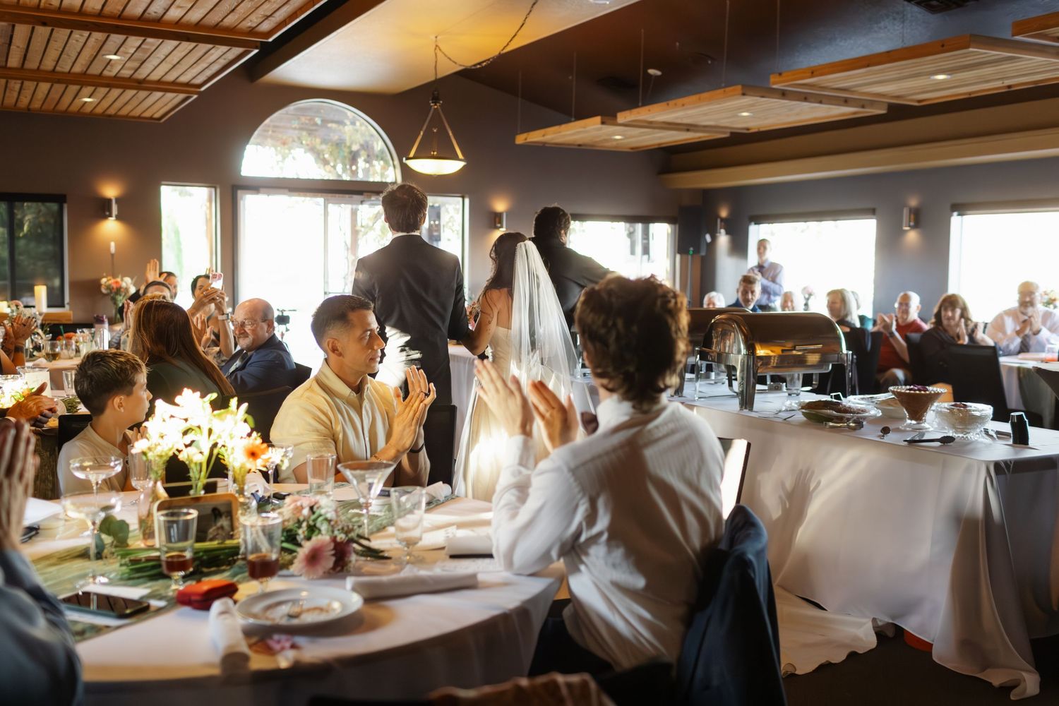 Bride and groom celebrate at elegant wedding reception dinner with guests seated at white-clothed tables in a restaurant.