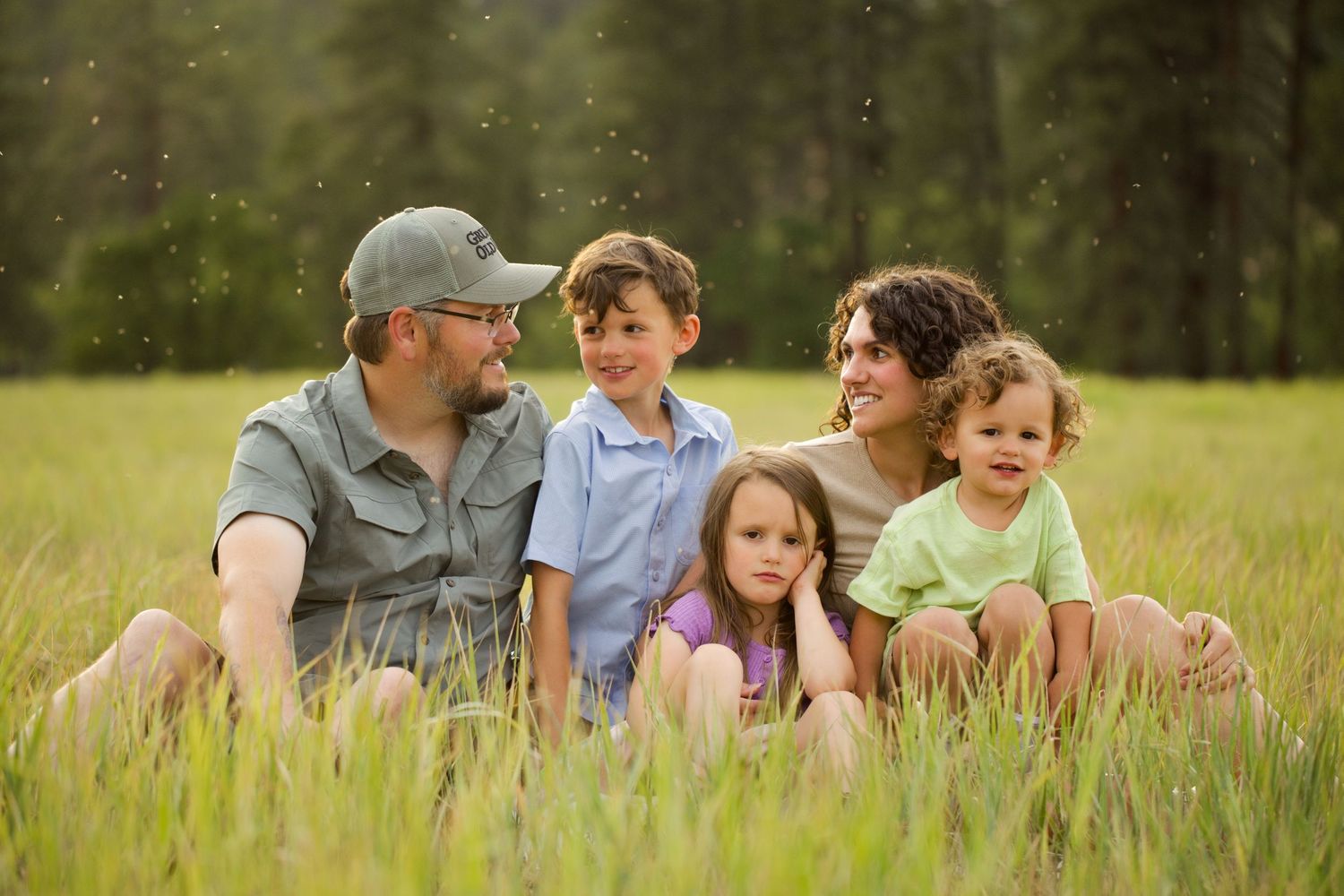 Family of five sitting together in a lush green meadow with tall grass and pine trees in the background.