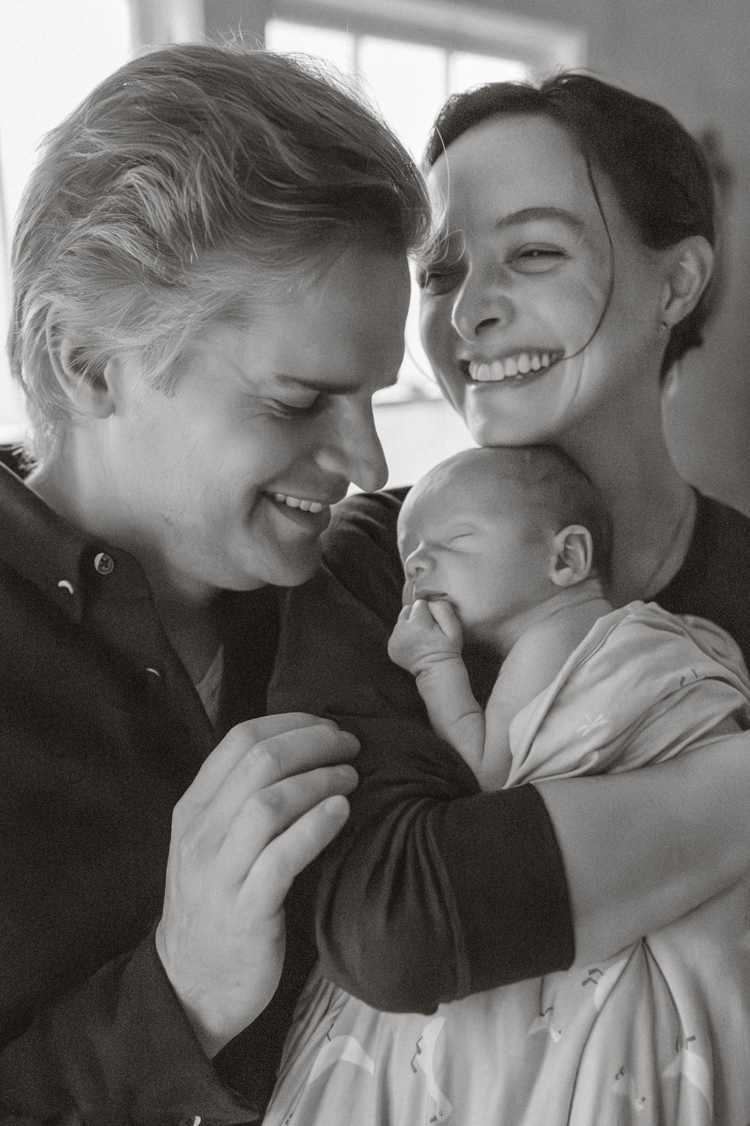 Black and white photo of two smiling parents lovingly gazing at their newborn baby.